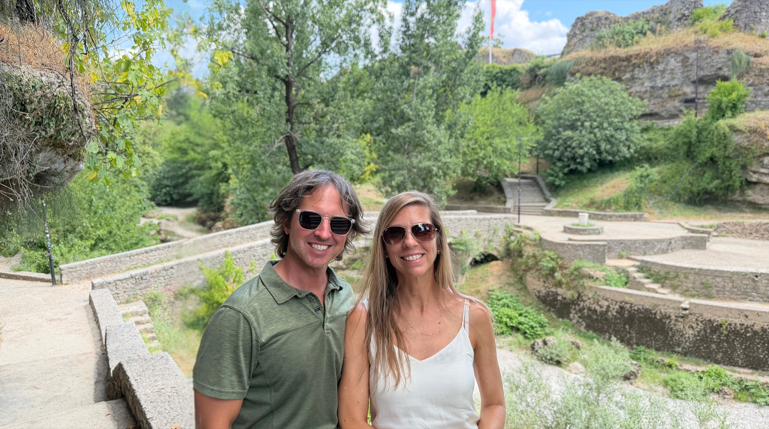 Sam and Stephanie by the Old Ribnica Bridge in Podgorica with stone ruins on the hill behind them
