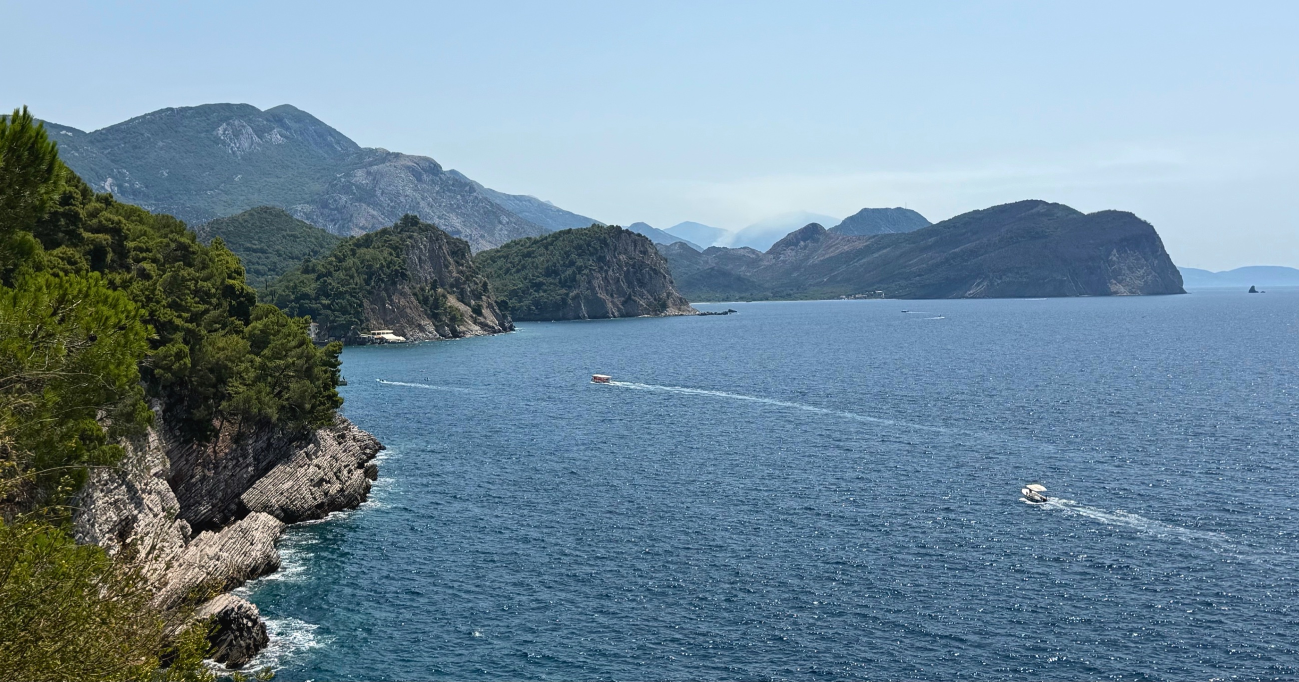 The Montenegrin coast north of Petrovac, with layered limestone cliffs, forested hills, and boats crossing the Adriatic.