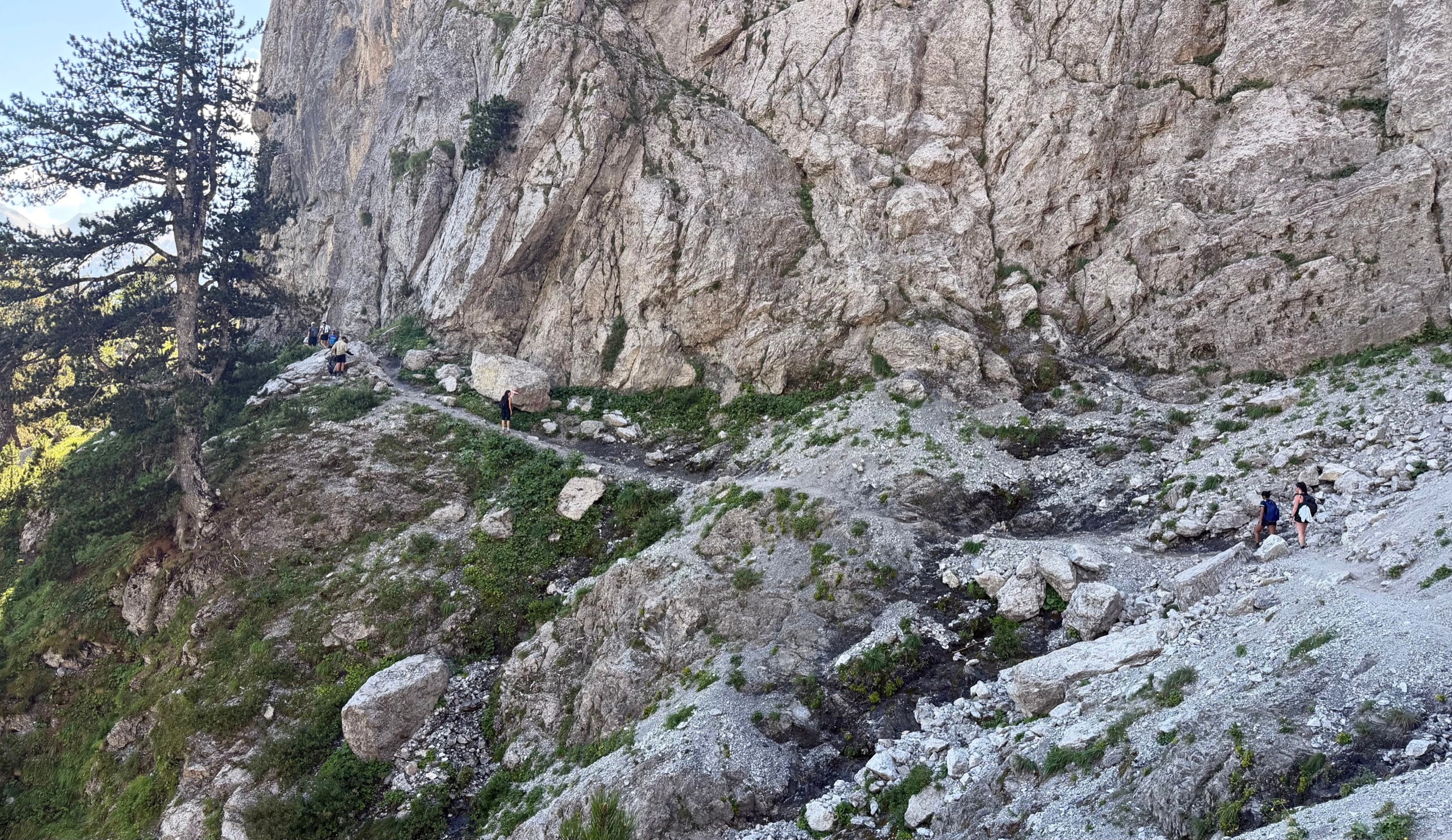 Hikers crossing a rocky trail beneath high limestone walls on the route toward Valbona Pass.