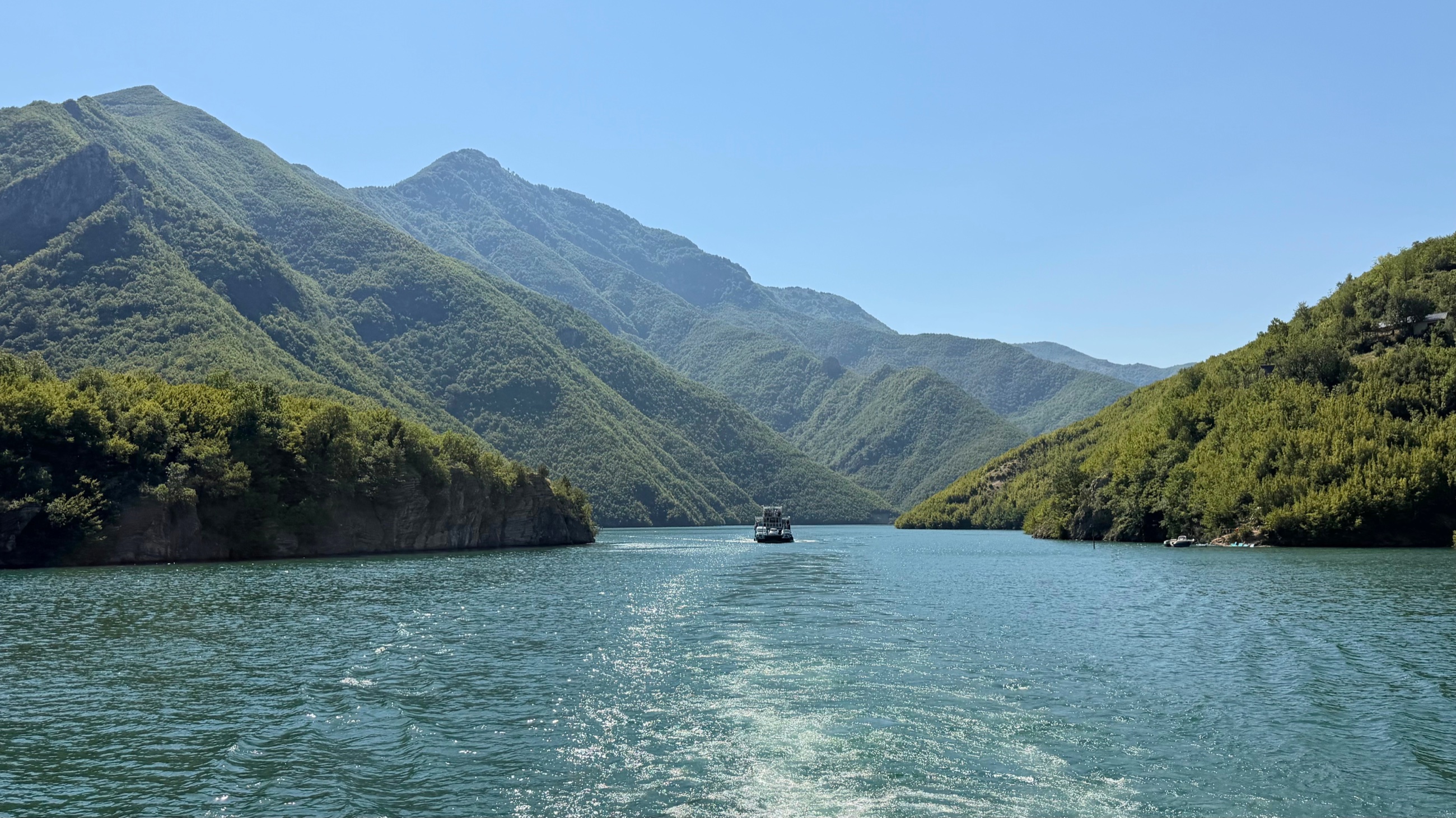 A boat ahead on the turquoise water of Komani Lake, surrounded by green mountains.