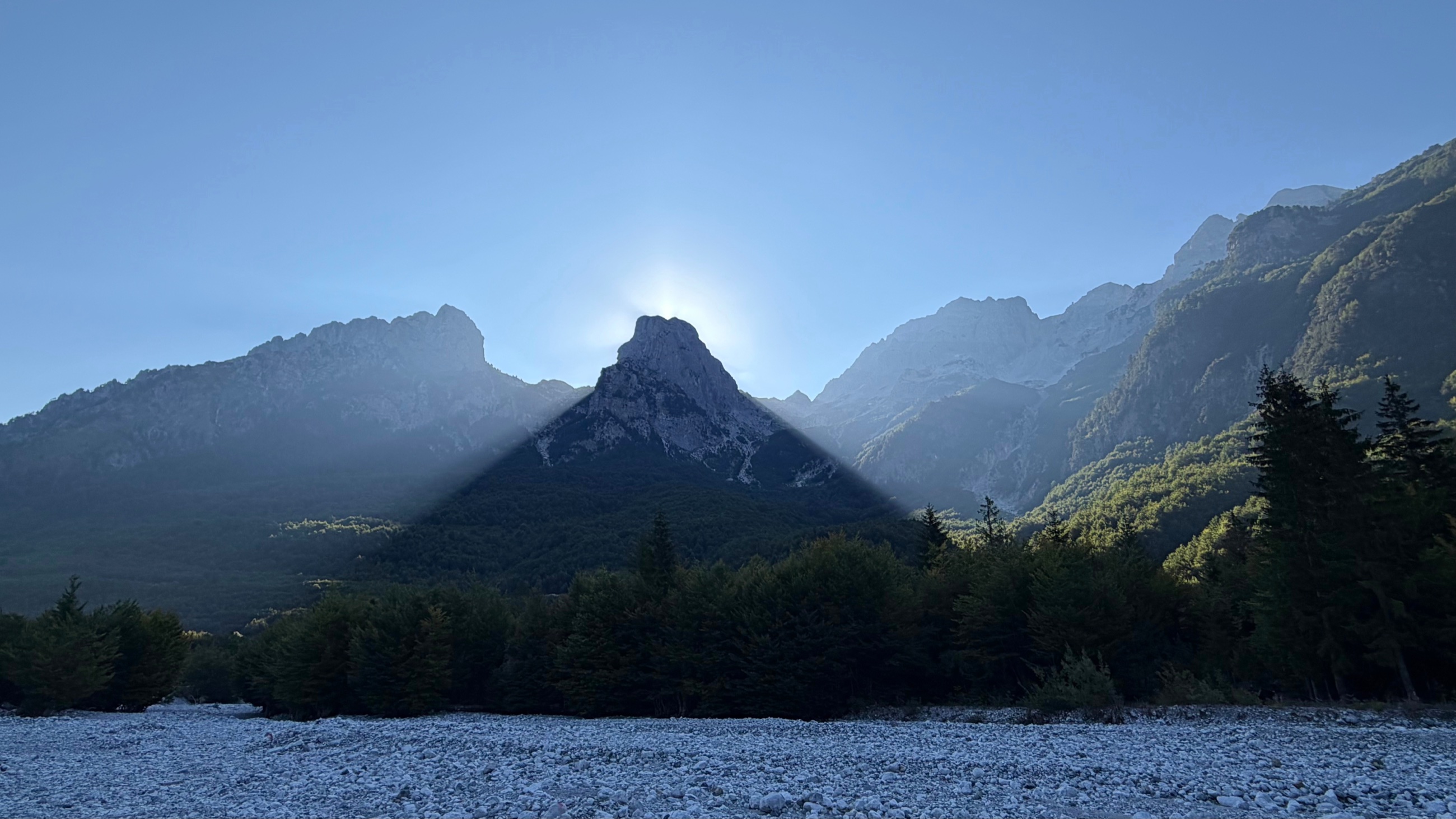 Morning sun breaking over the mountains above the pale riverbed in Valbona Valley.