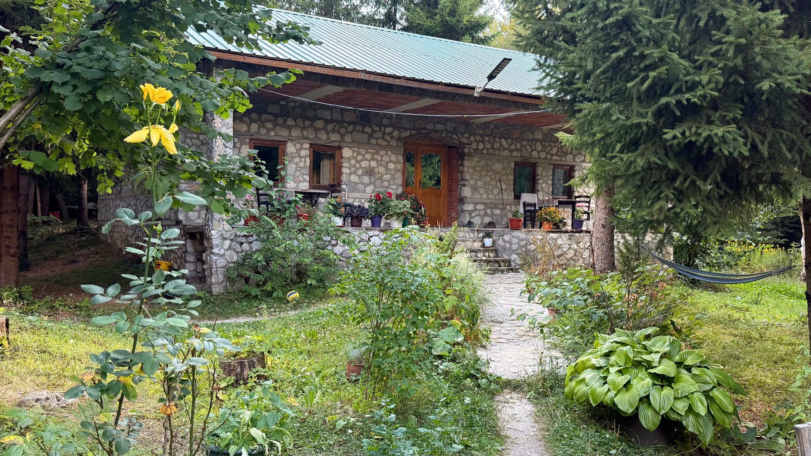 Stone guesthouse in Valbona surrounded by gardens, trees, and a hammock.