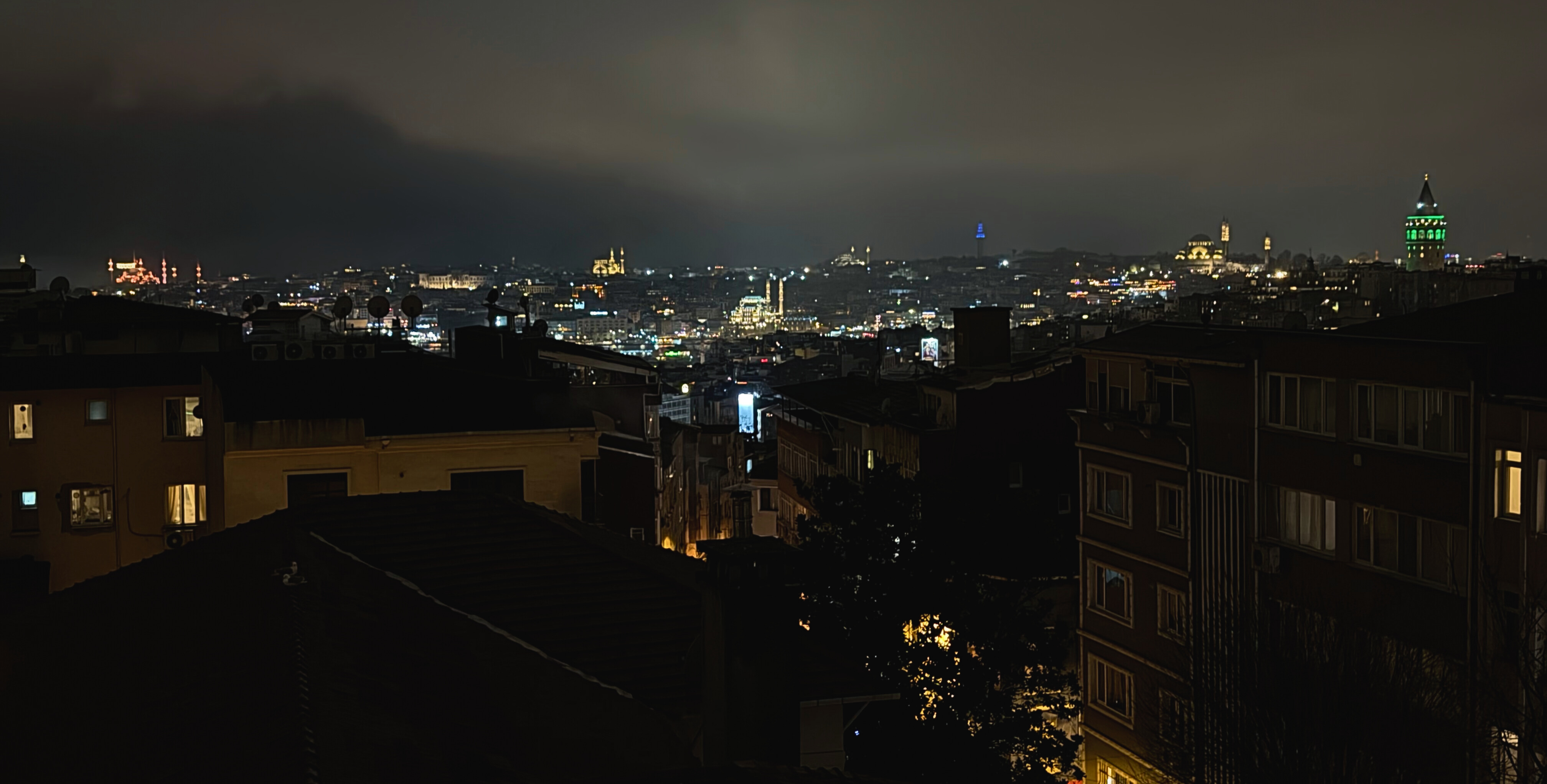 Night view over Istanbul rooftops with Galata Tower and mosque lights visible across the city.
