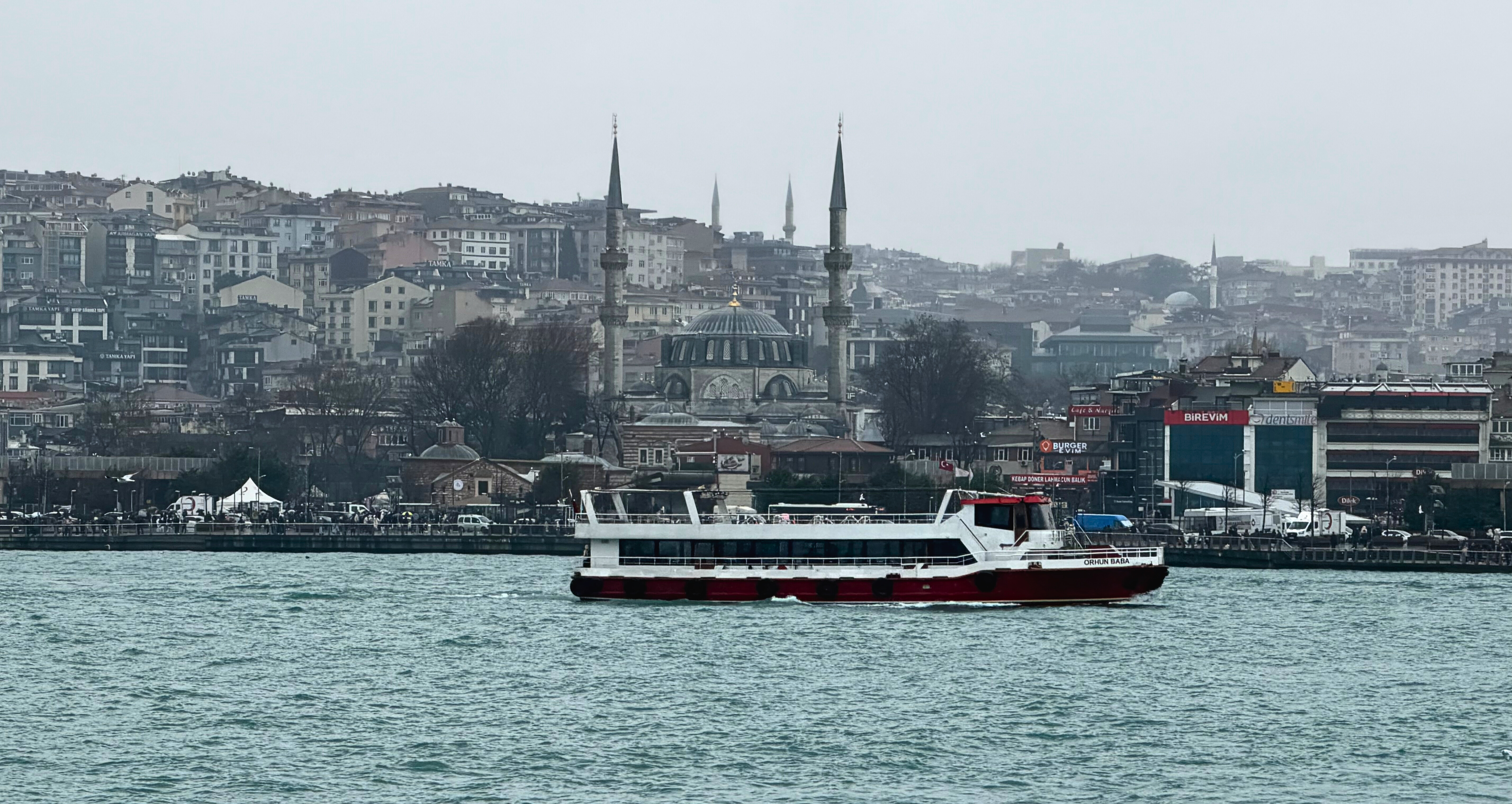 A passenger ferry crossing the water in Istanbul with mosques and hillside buildings behind it.
