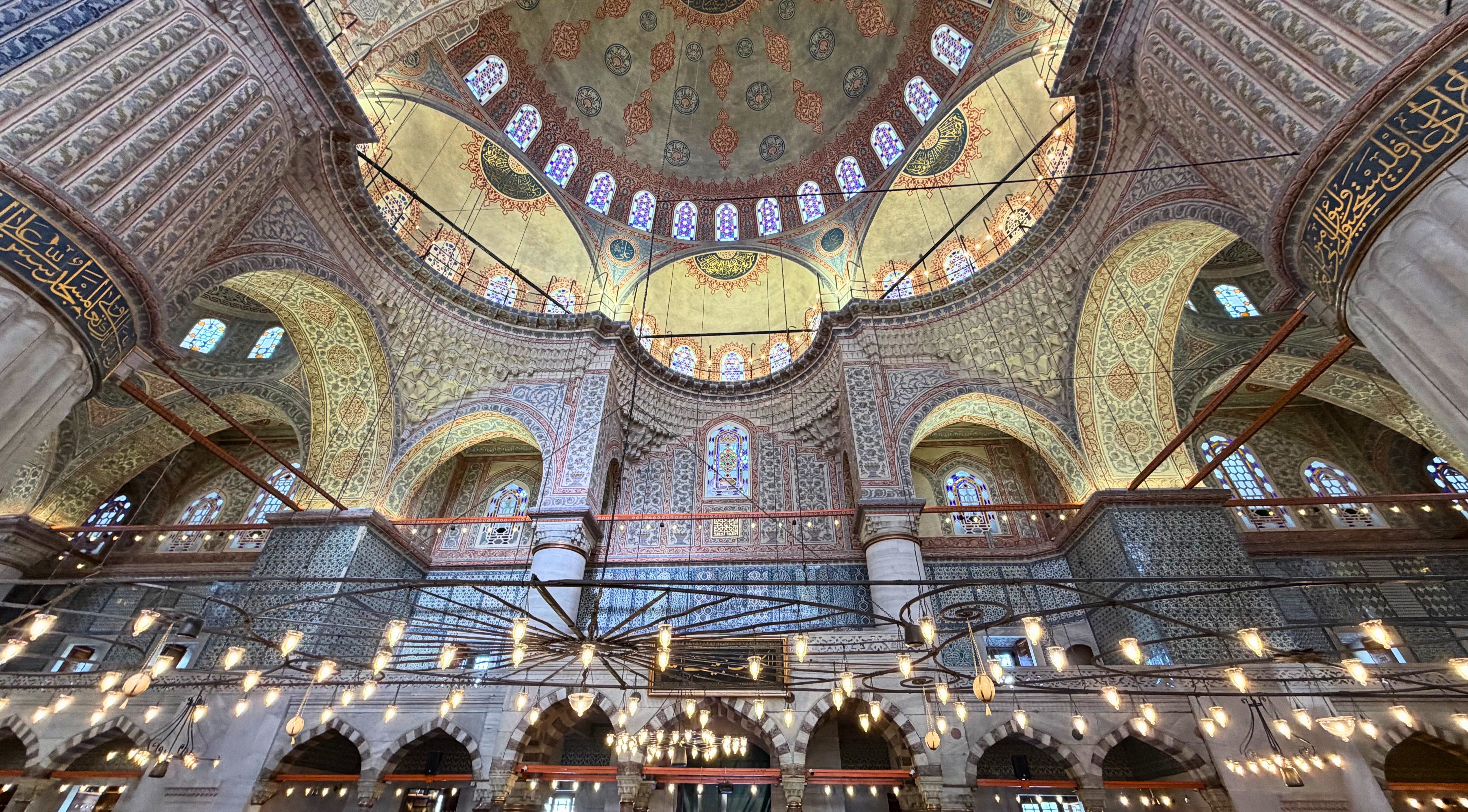 The interior of the Blue Mosque with patterned domes, arches, hanging lights, and blue İznik tiles.