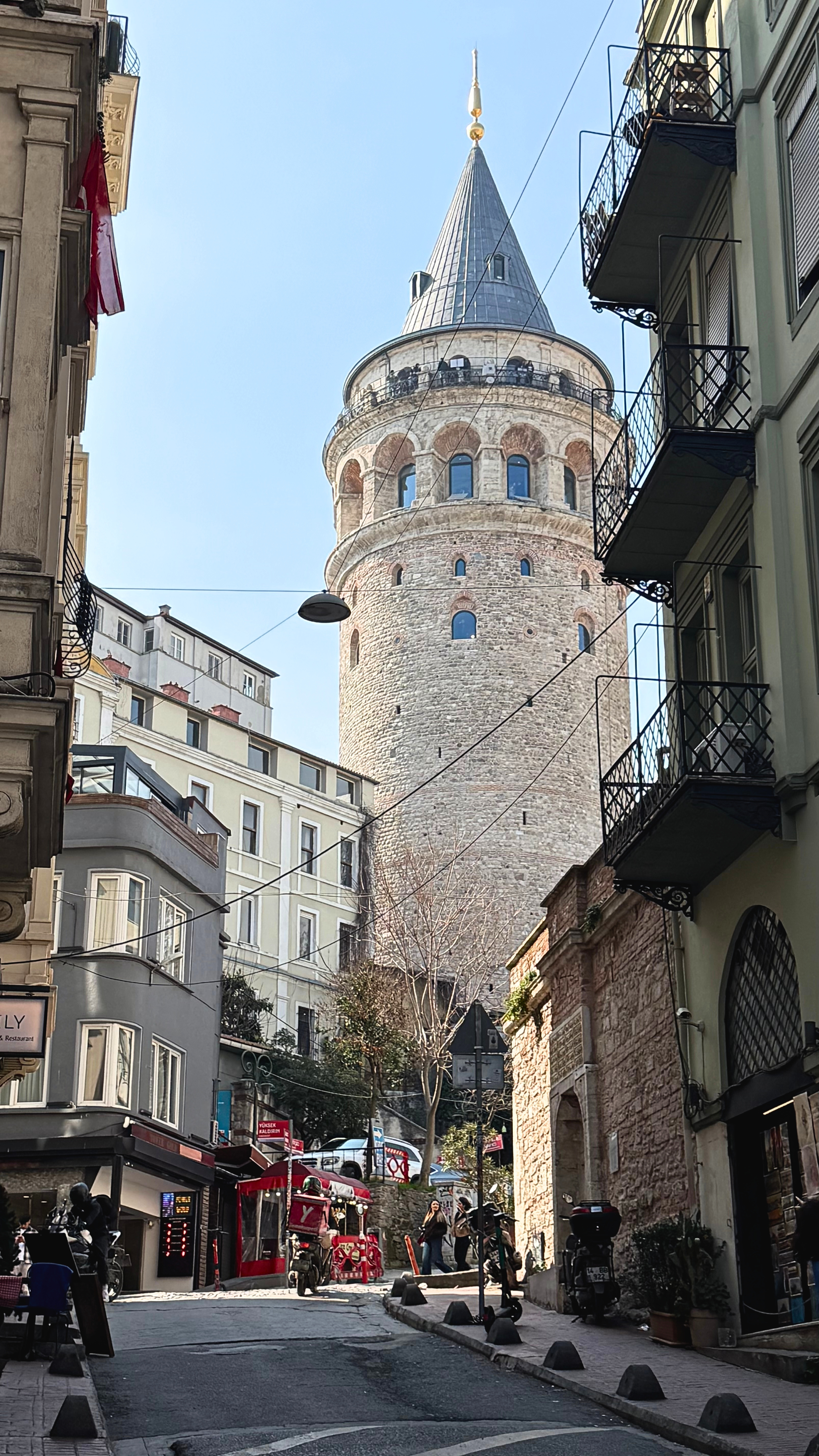 Galata Tower rising above a narrow street lined with buildings, balconies, and pedestrians.