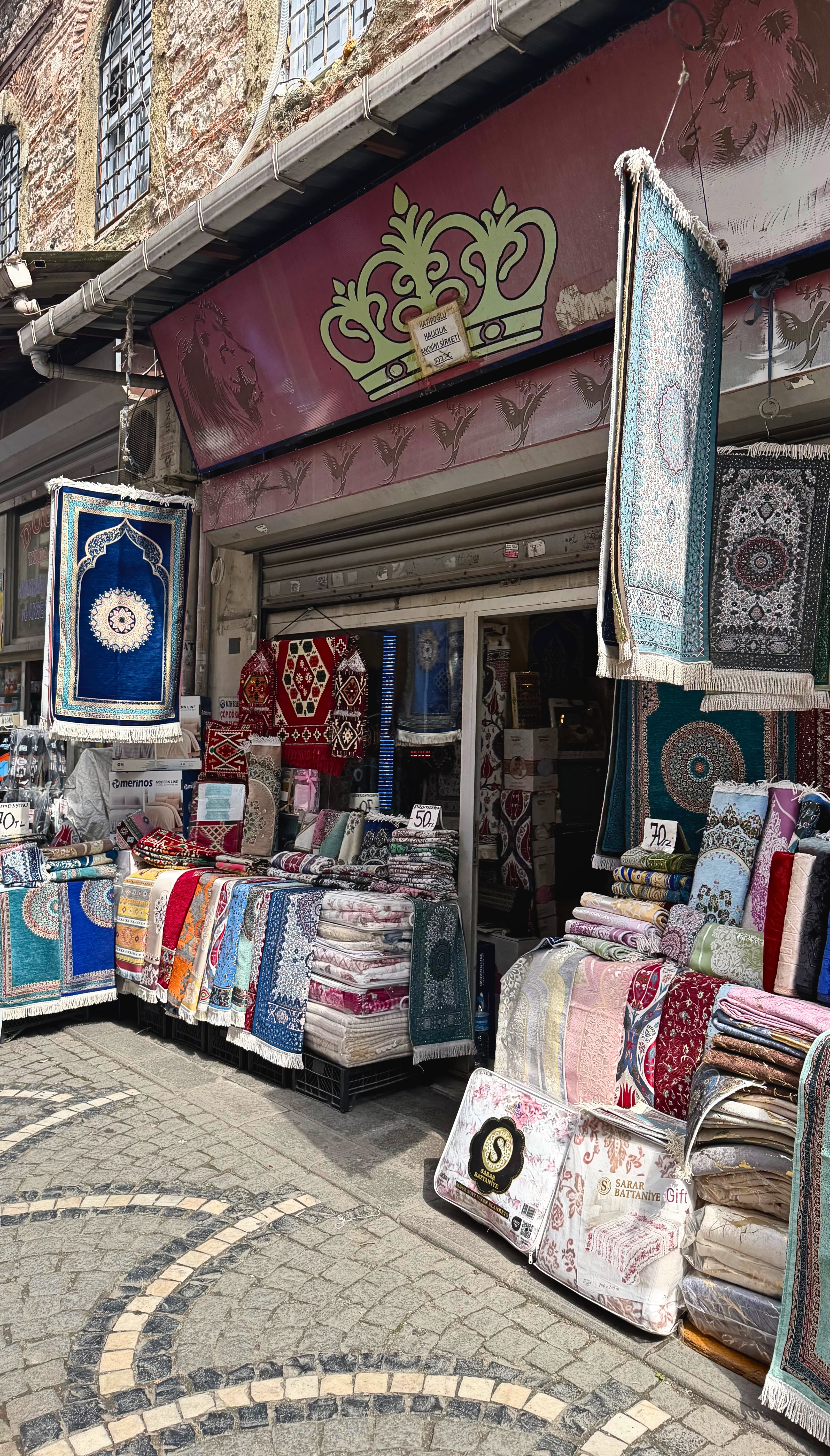 A market storefront displaying prayer rugs, blankets, patterned textiles, and folded goods.