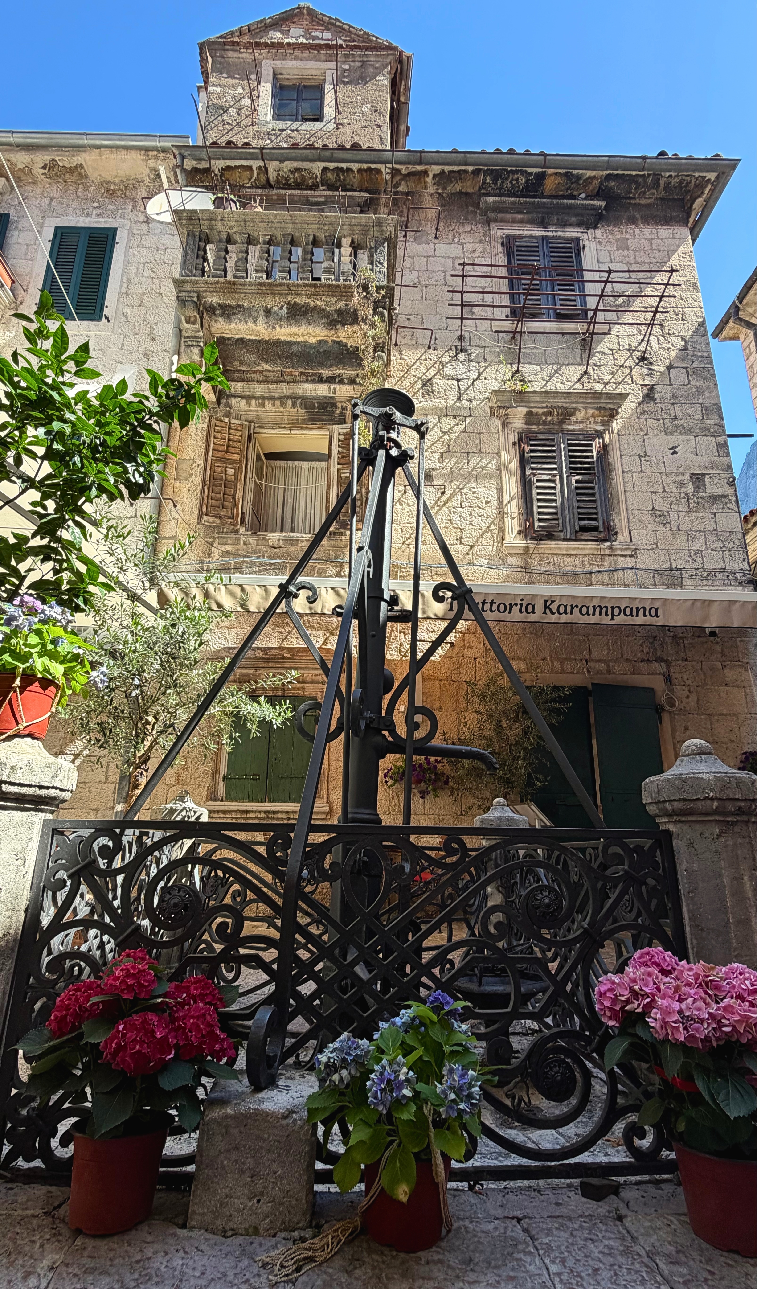 The Karampana wellhead among stone walls, shutters, and flowers in the Old Town.