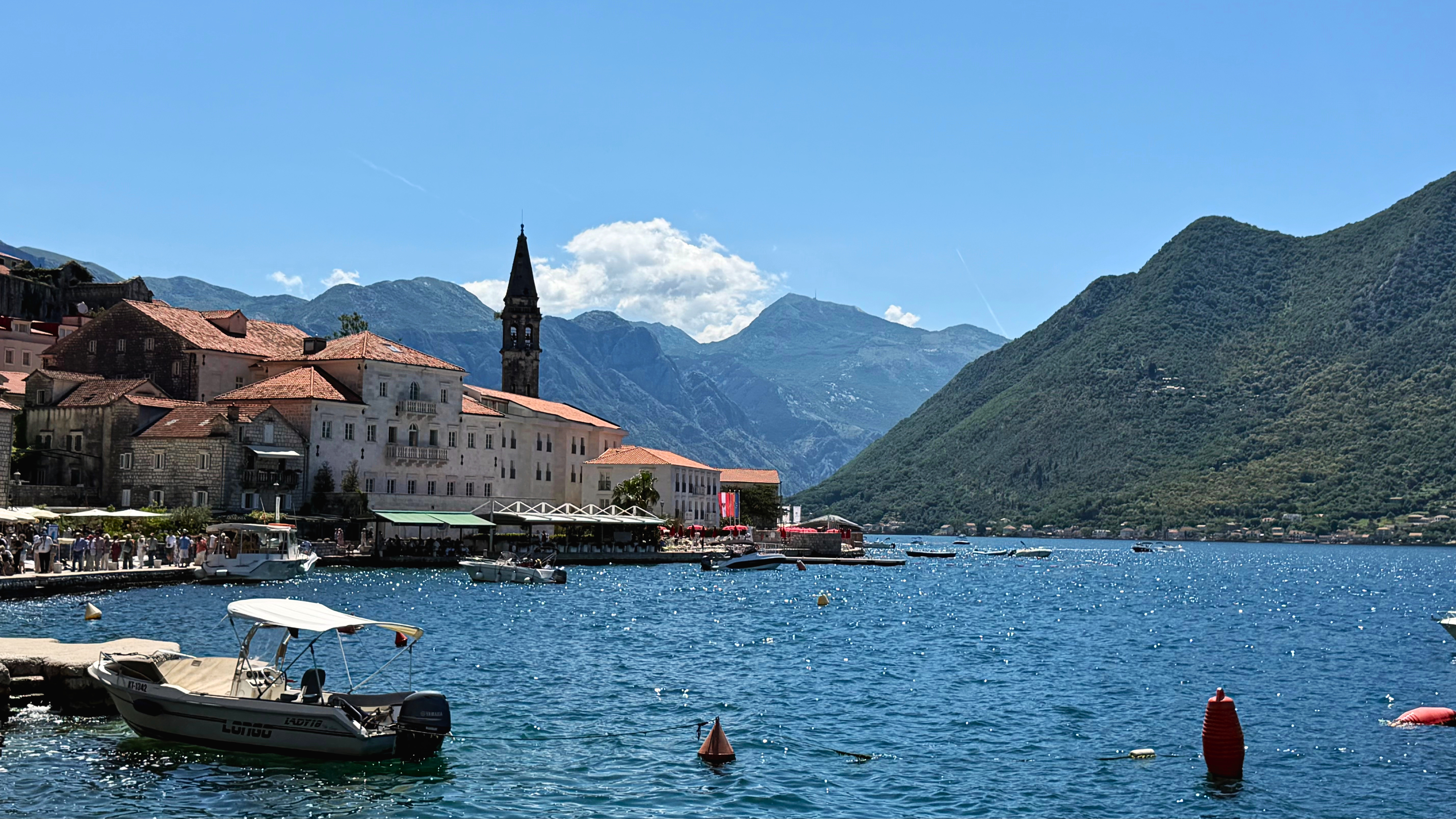 Perast from the waterline, with stone houses, church towers, boats, and mountains along the bay.