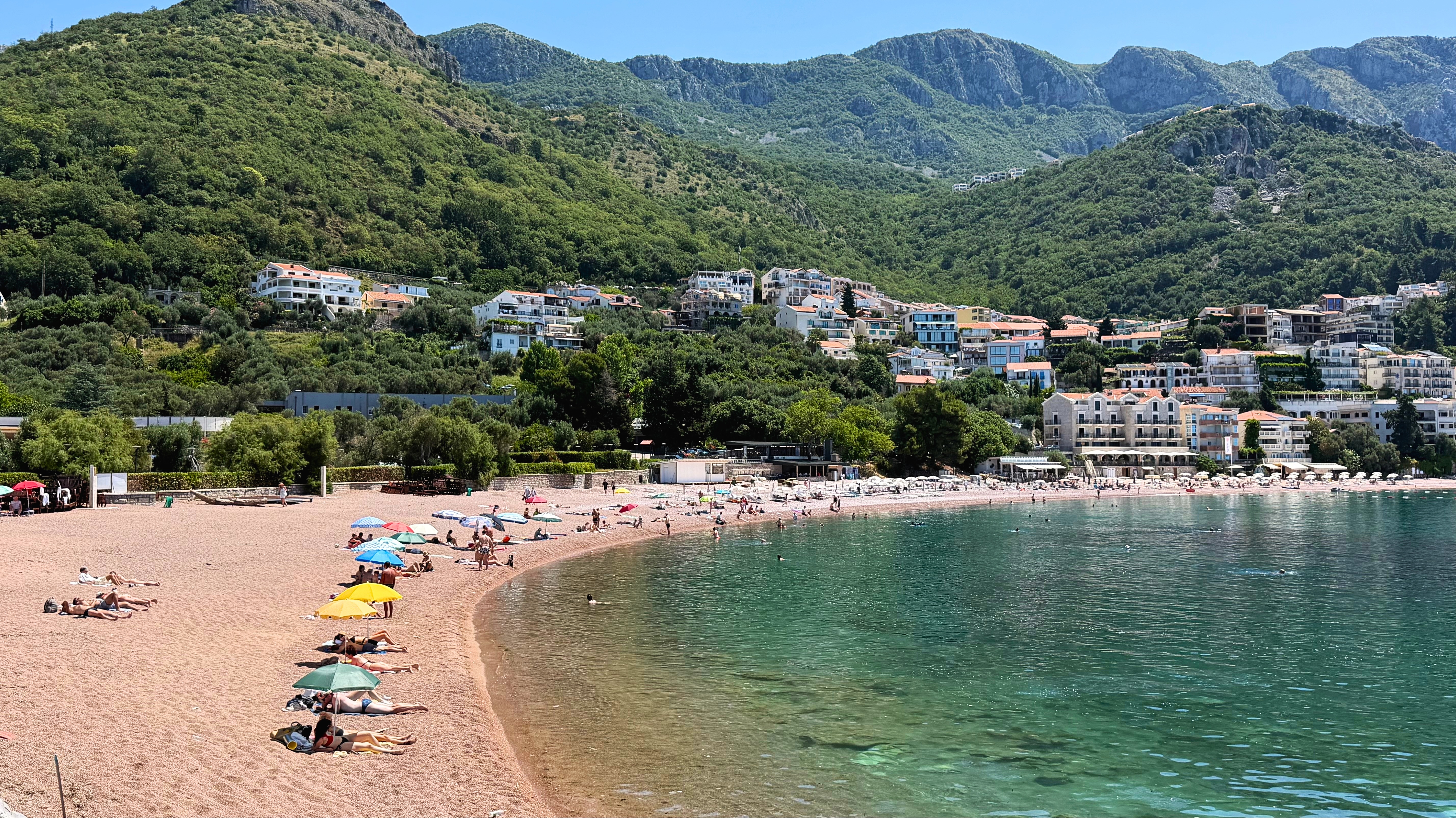 The public beach below Sveti Stefan, with clear water, pink pebbles, and mountains behind the shore.
