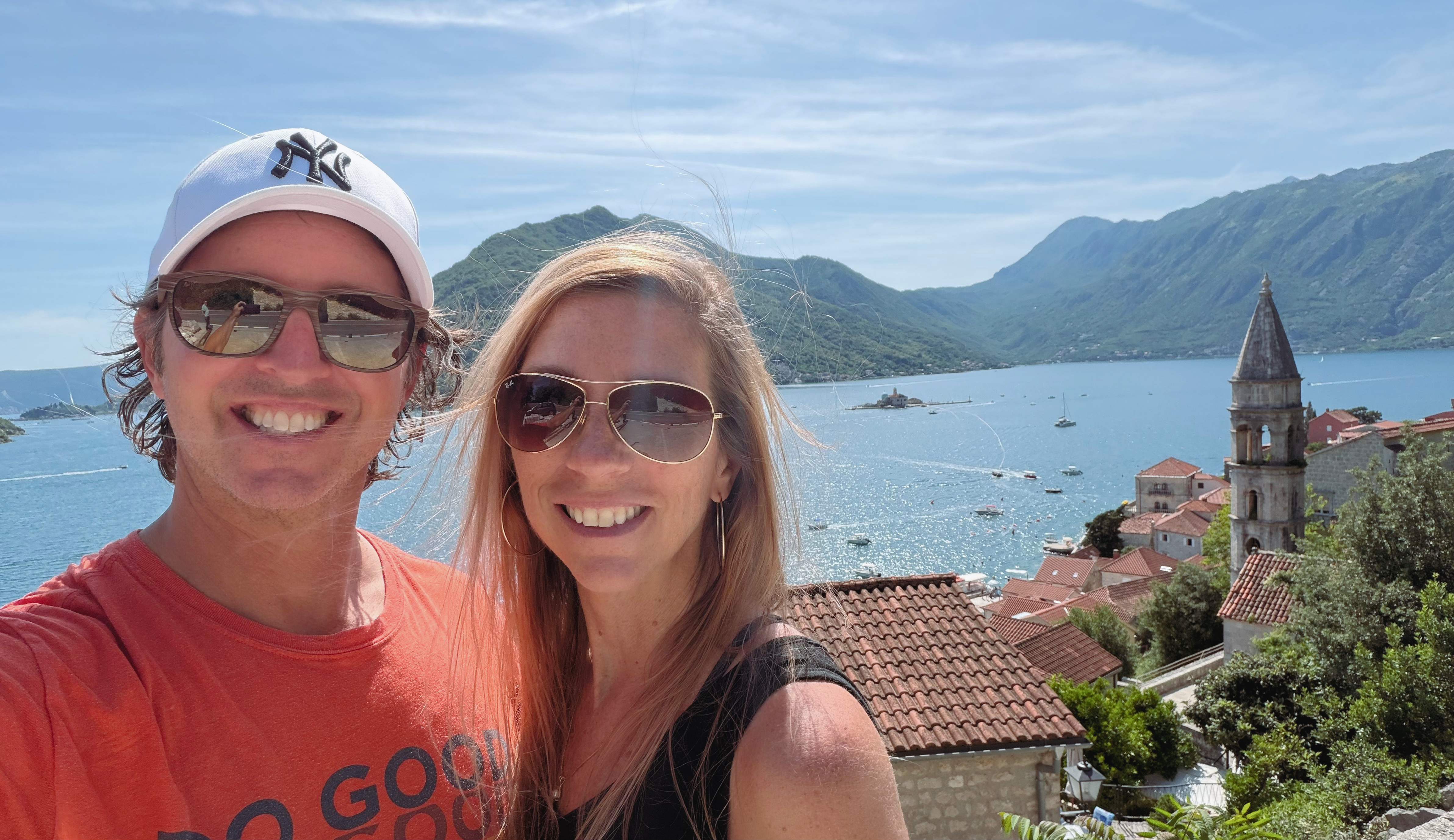 Sam and Stephanie above Perast, with the bay, rooftops, bell tower, and island churches behind them.