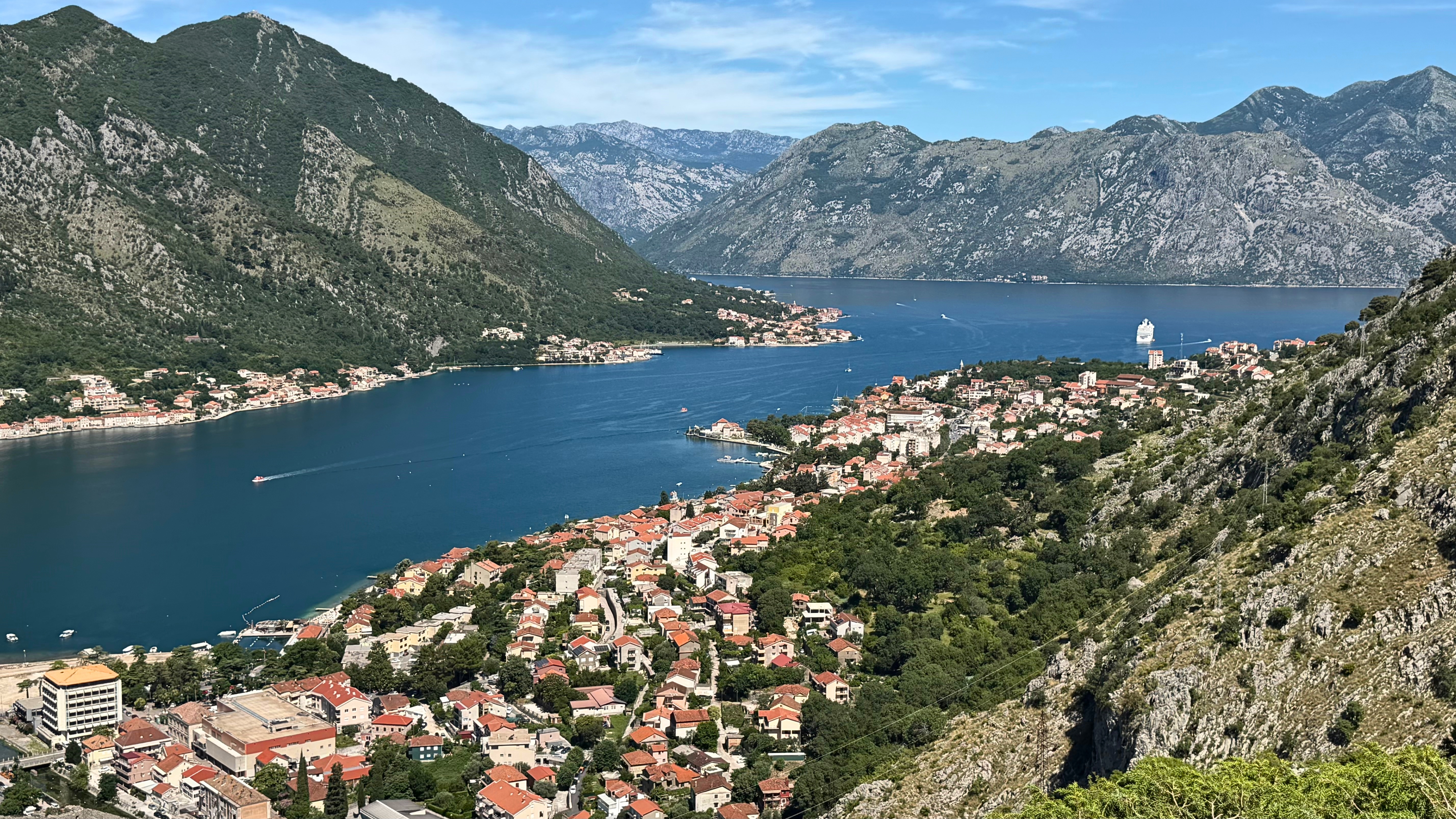 The Bay of Kotor from above, with Kotor and Dobrota merged into the wider shape of the bay, red roofs, blue water, steep mountains, and one cruise ship