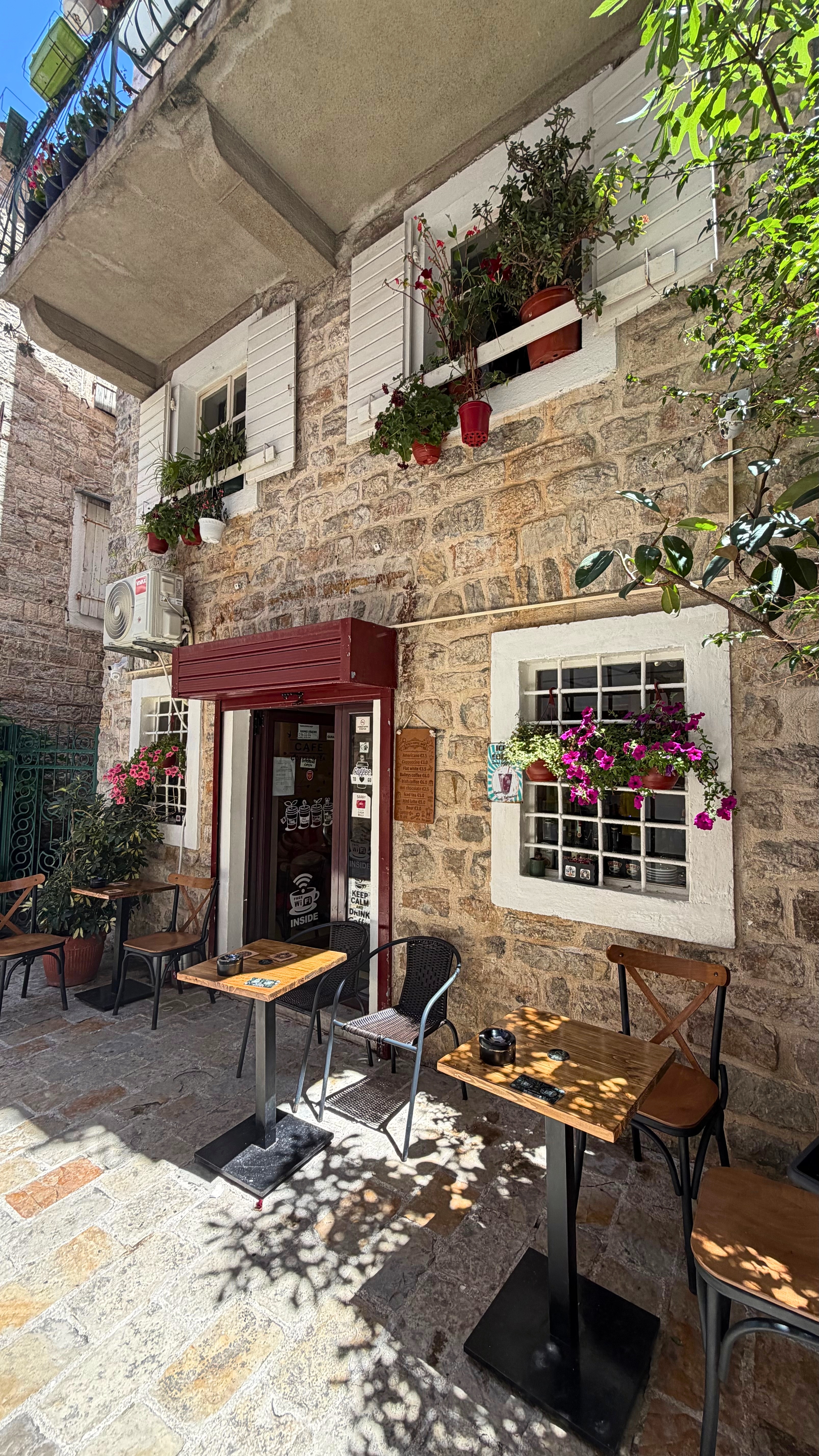 A quiet courtyard at North Coffee in Kotor Old Town, with stone walls, shutters, flowers, and shaded tables