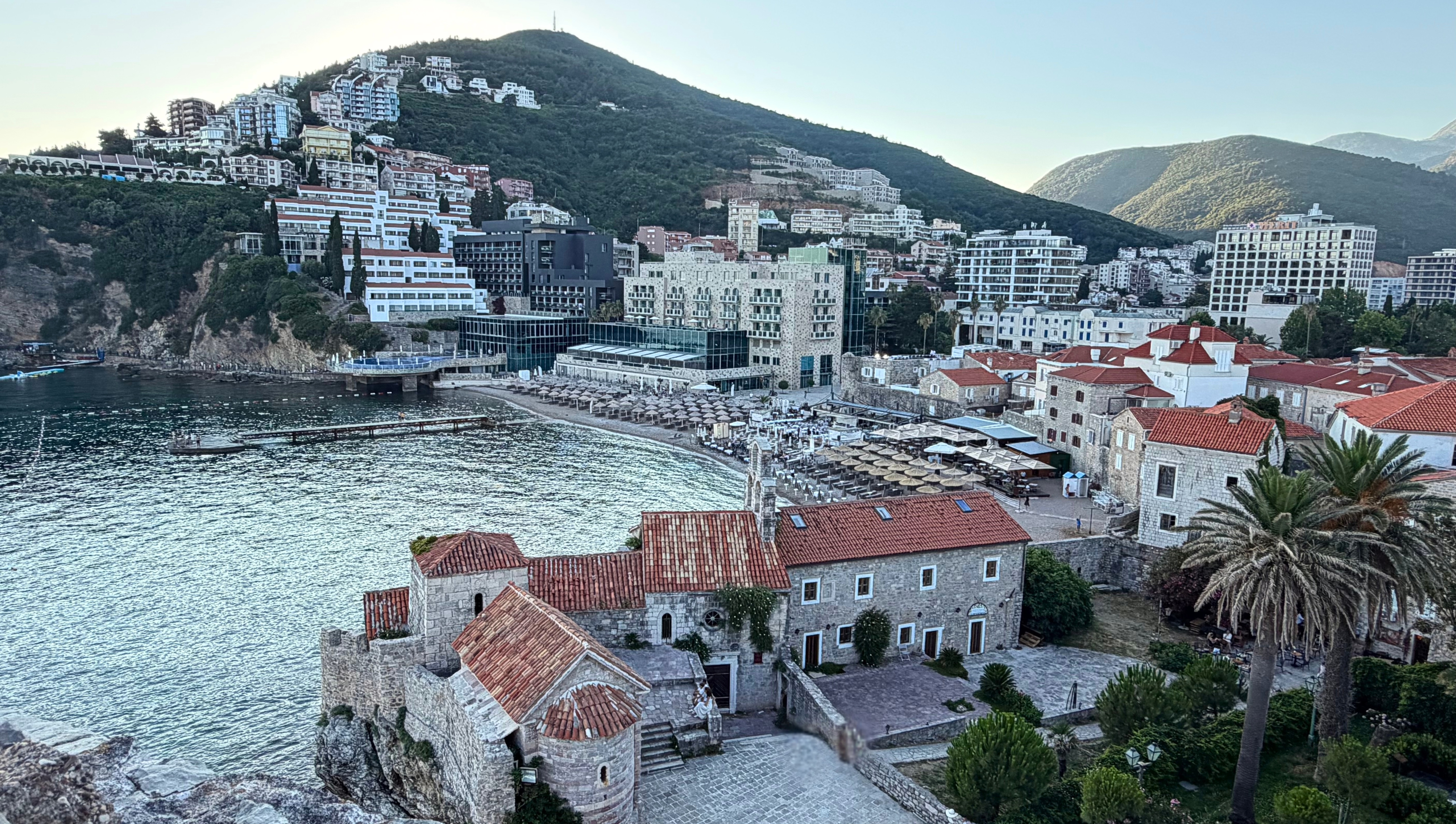 Budva from the Citadel, old stone, beach umbrellas, modern hotels, and hillside apartments pressed together along the coast