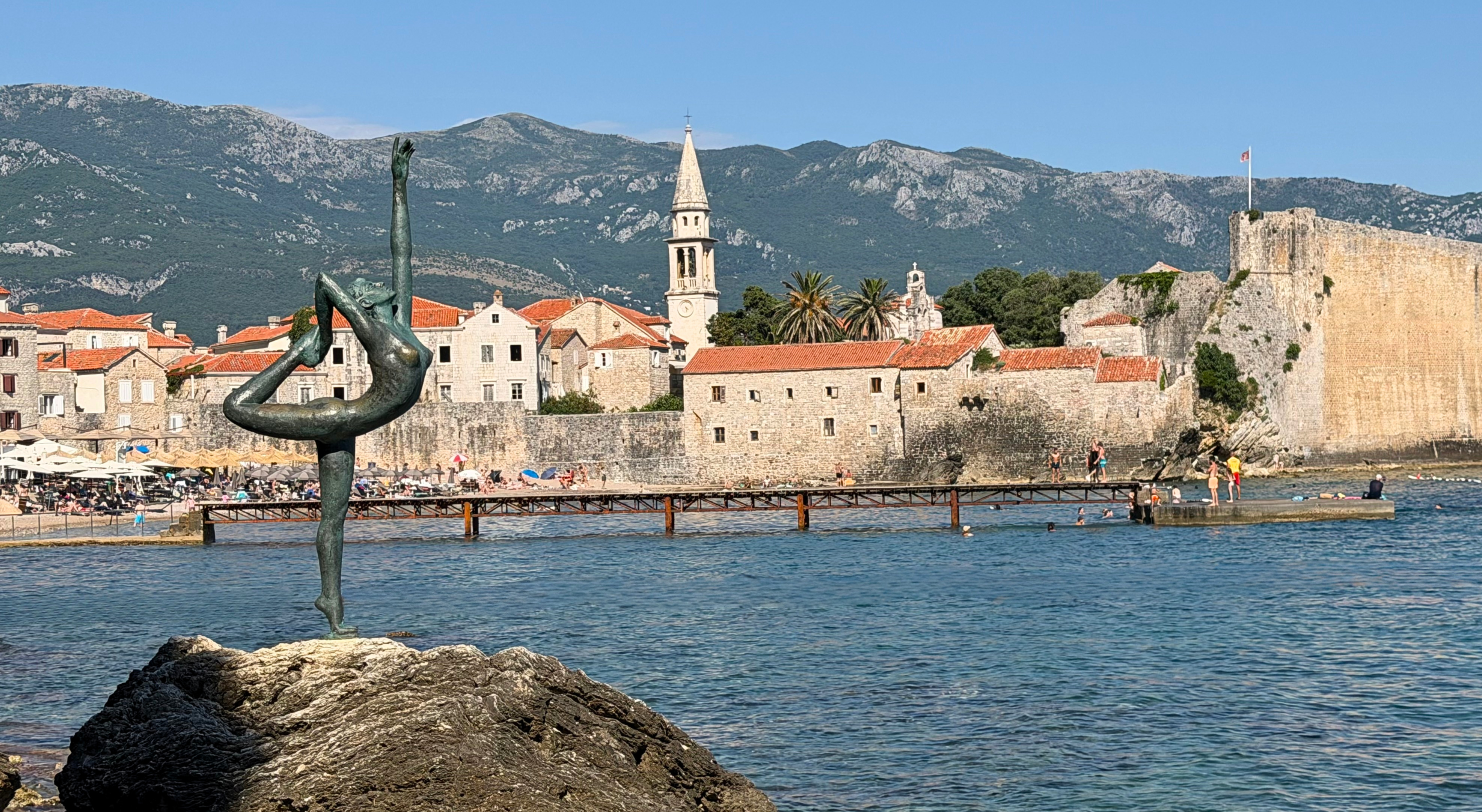 The Ballet Dancer statue in Budva, set before the old walls, the water, the church tower, and the mountains behind town