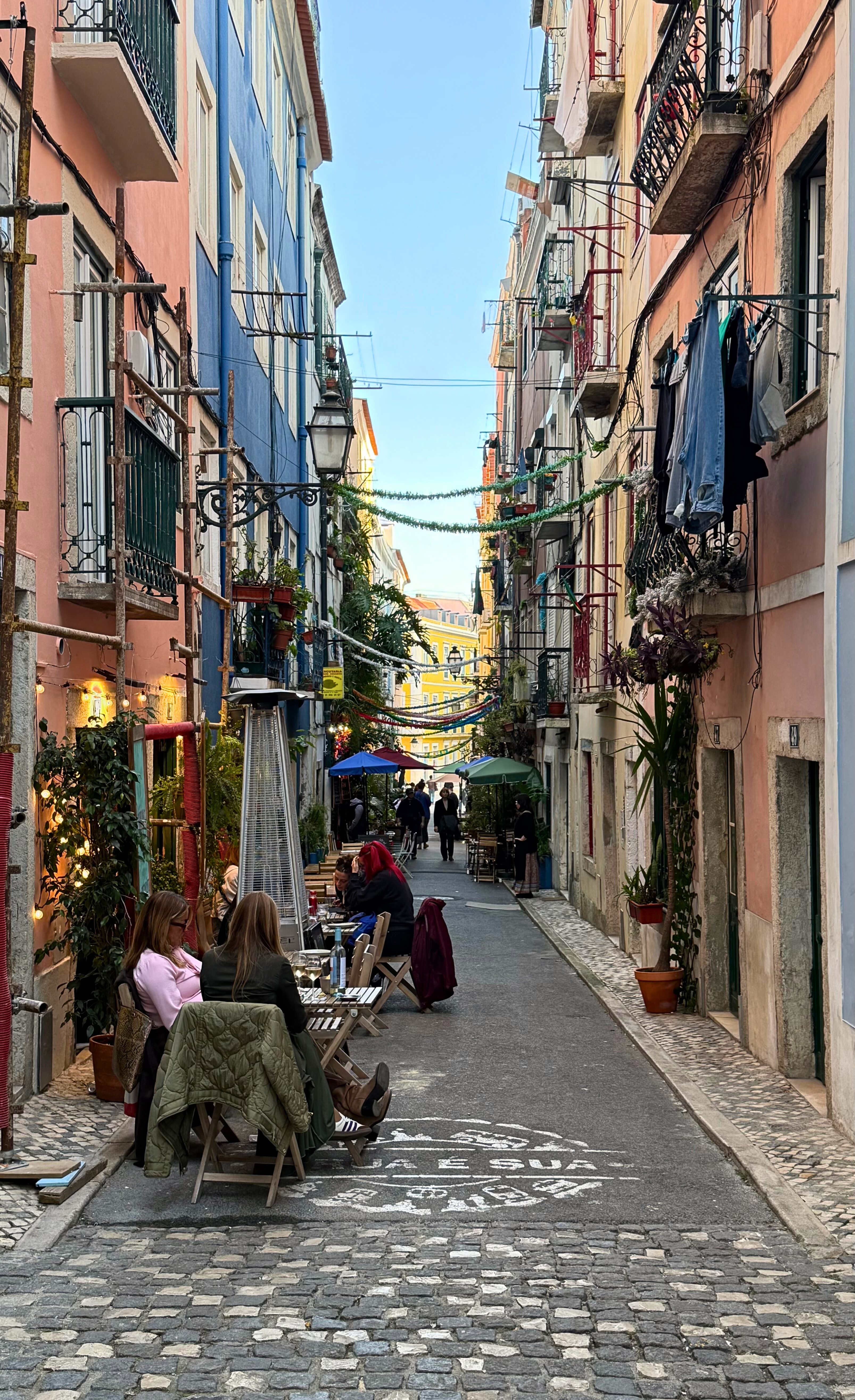 A narrow Lisbon street with green-painted buildings, balconies, and people gathered outside.