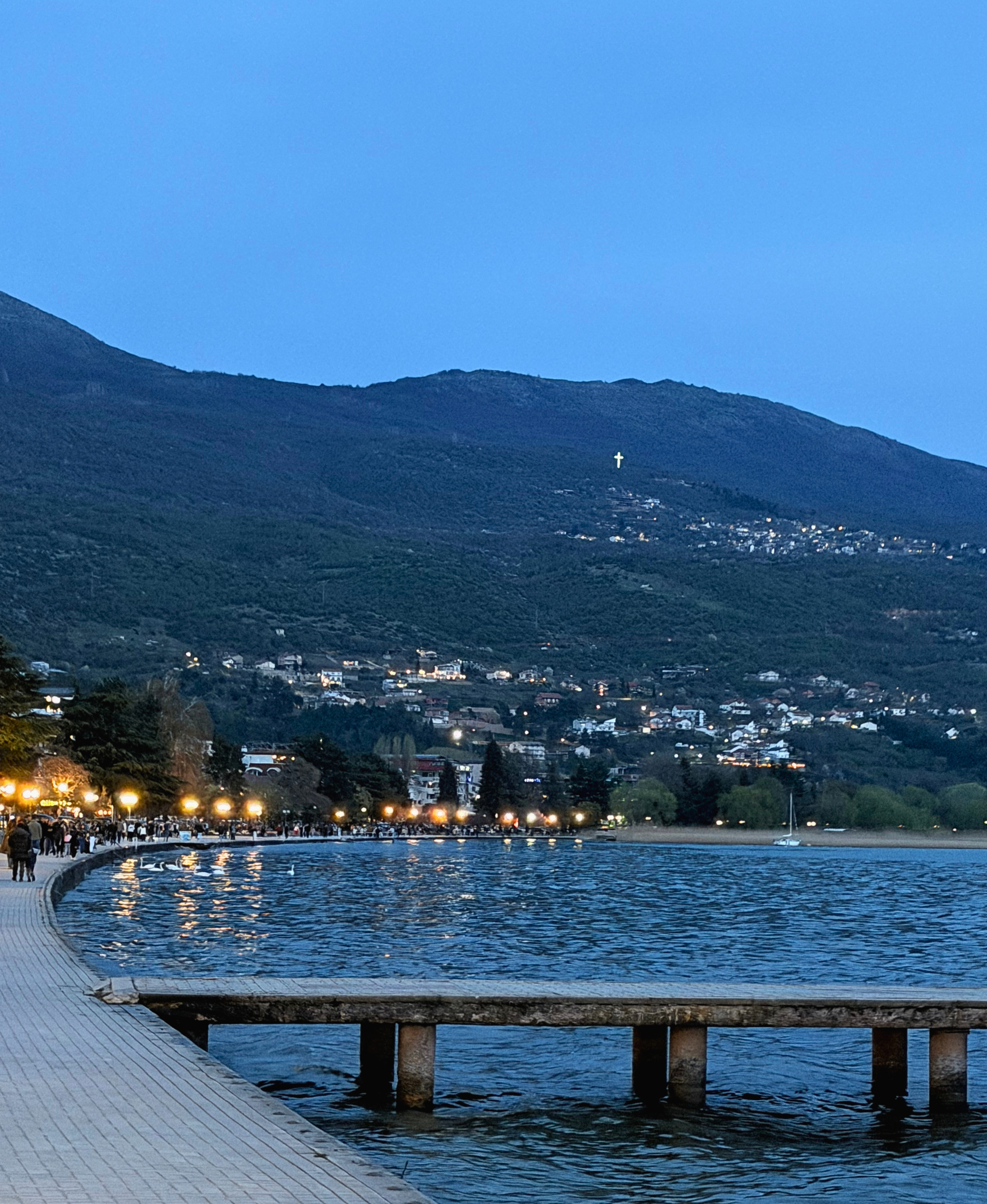 Ohrid promenade at dusk with a lit cross on the hillside above the lake.