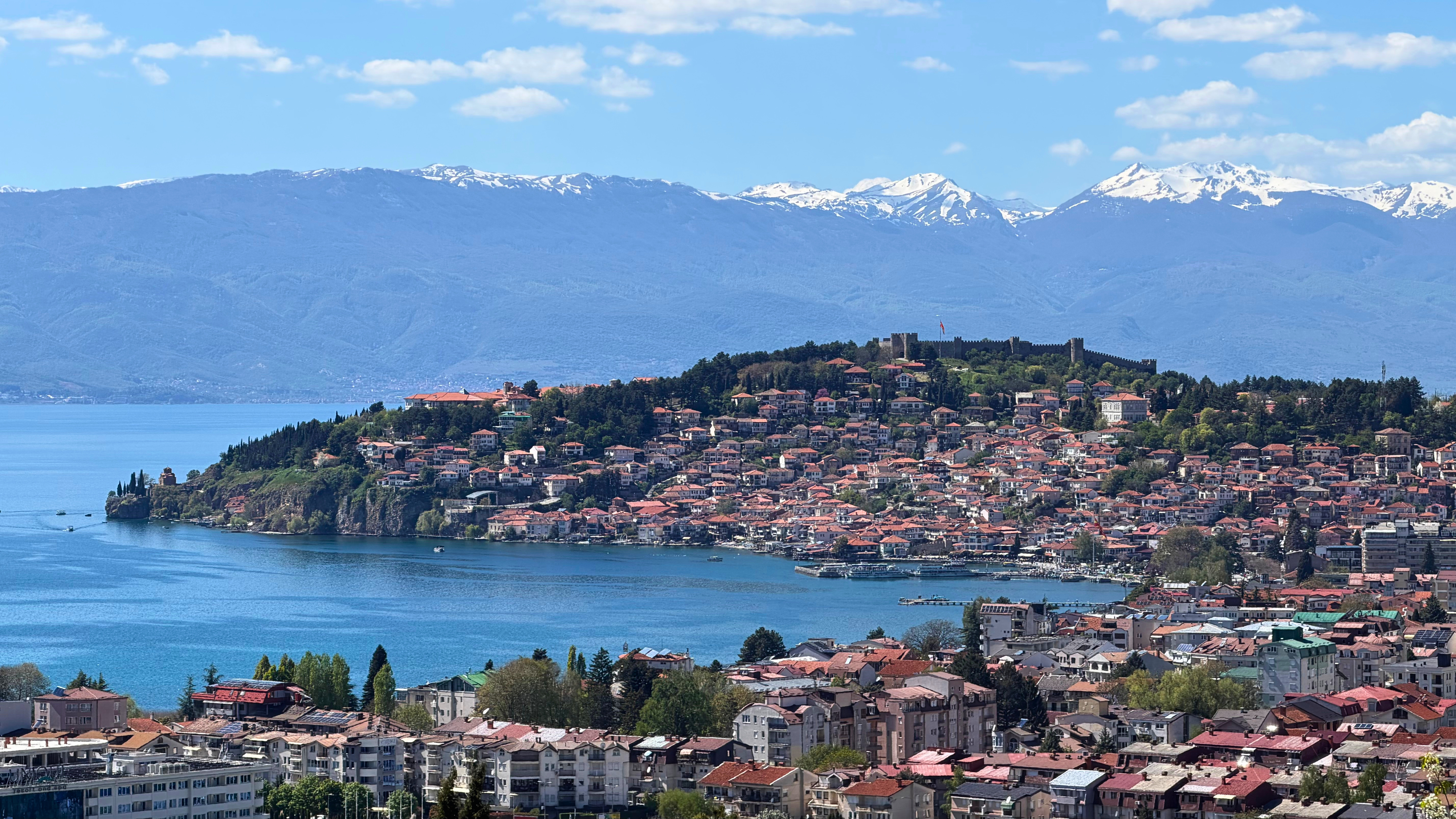 Ohrid seen from a hillside trail, with red rooftops, the old fortress, Lake Ohrid, and snow-capped mountains beyond.