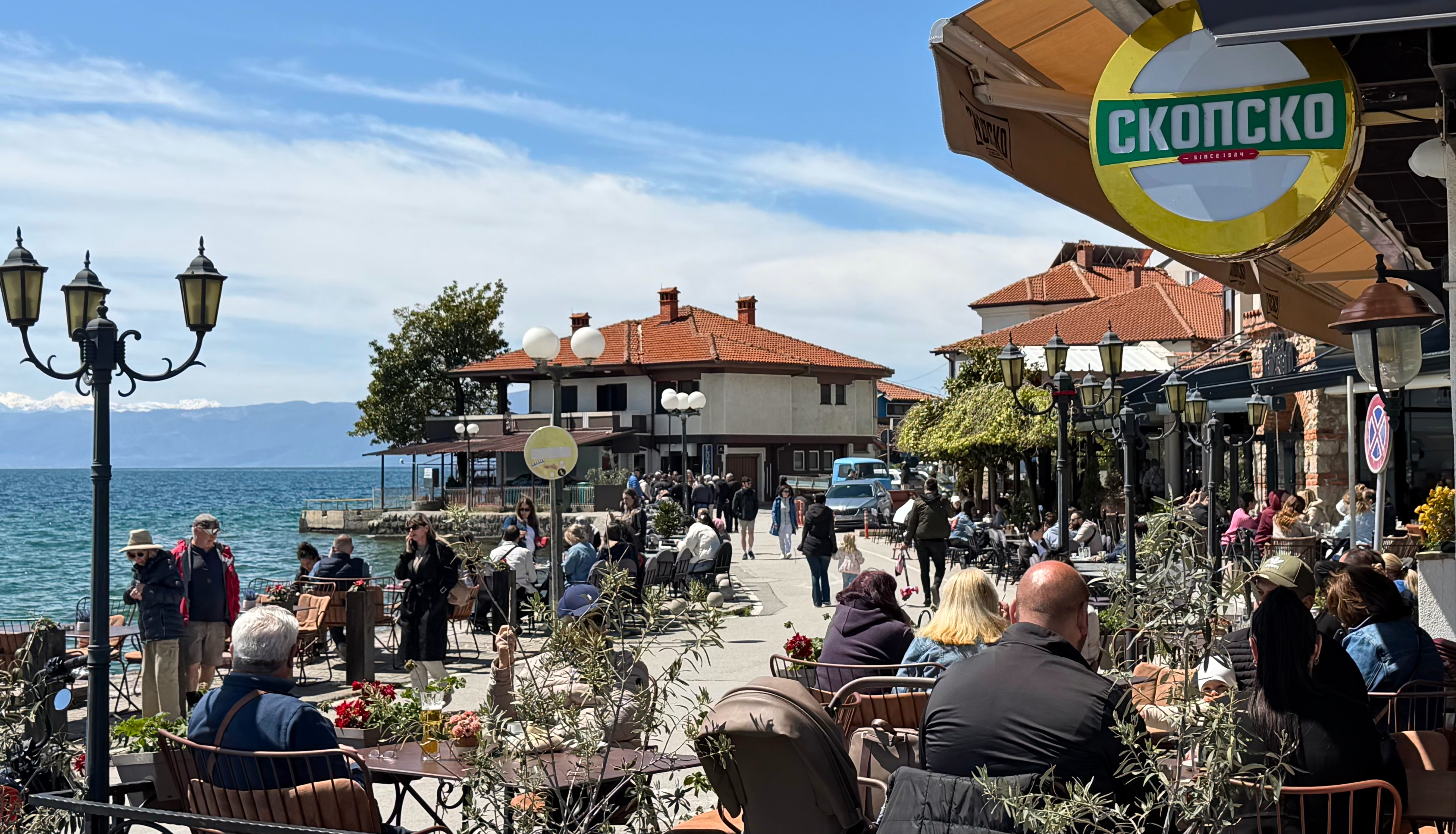 A busy sunny waterfront lane in Ohrid with cafés, people, lake views, and old town buildings.