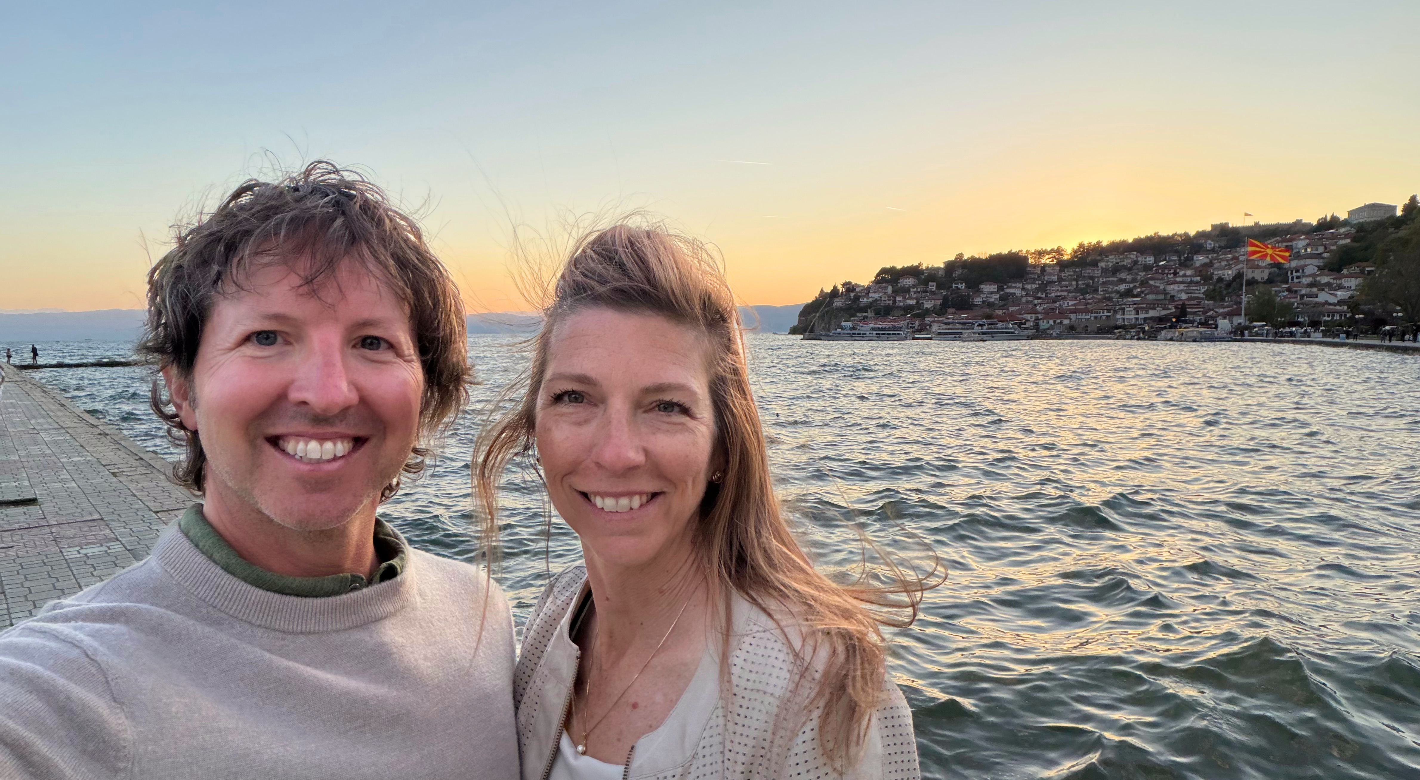 Sam and Stephanie standing on the Ohrid waterfront at sunset, with the lake, old town, and North Macedonian flag behind them.
