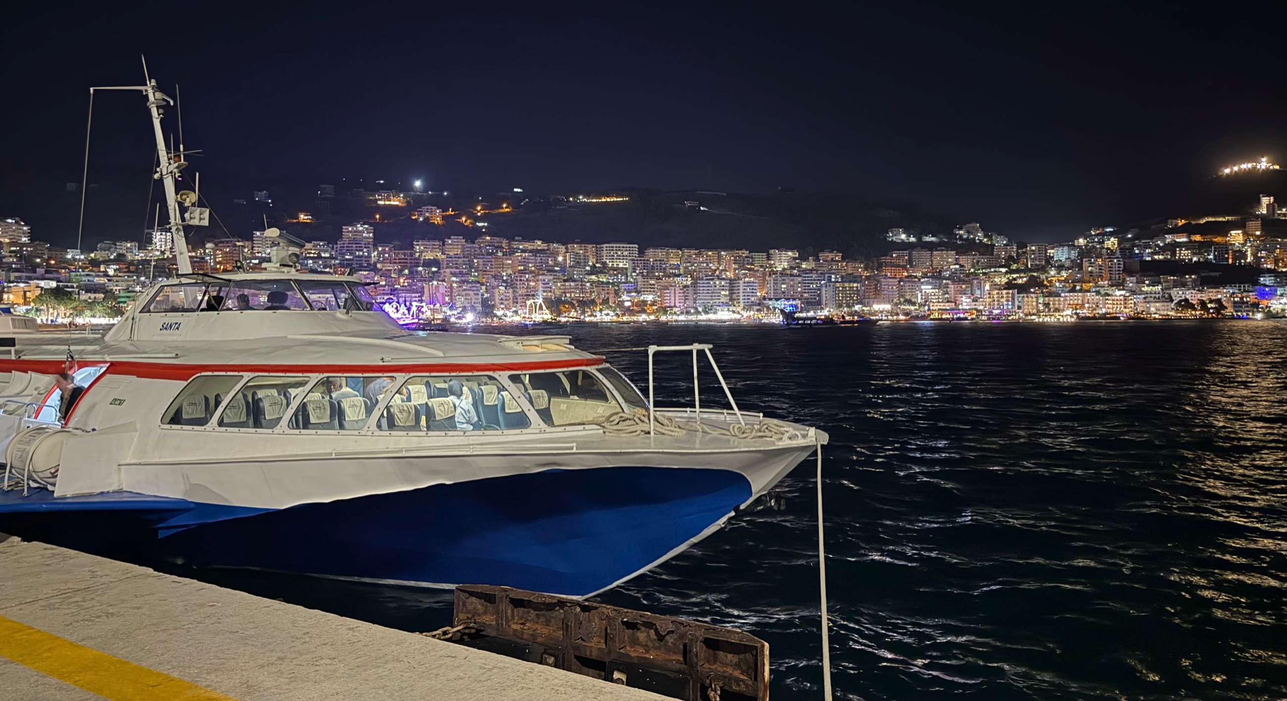 The Sarandë-Corfu hydrofoil docked at night along Sarandë's waterfront with the illuminated city across the bay.