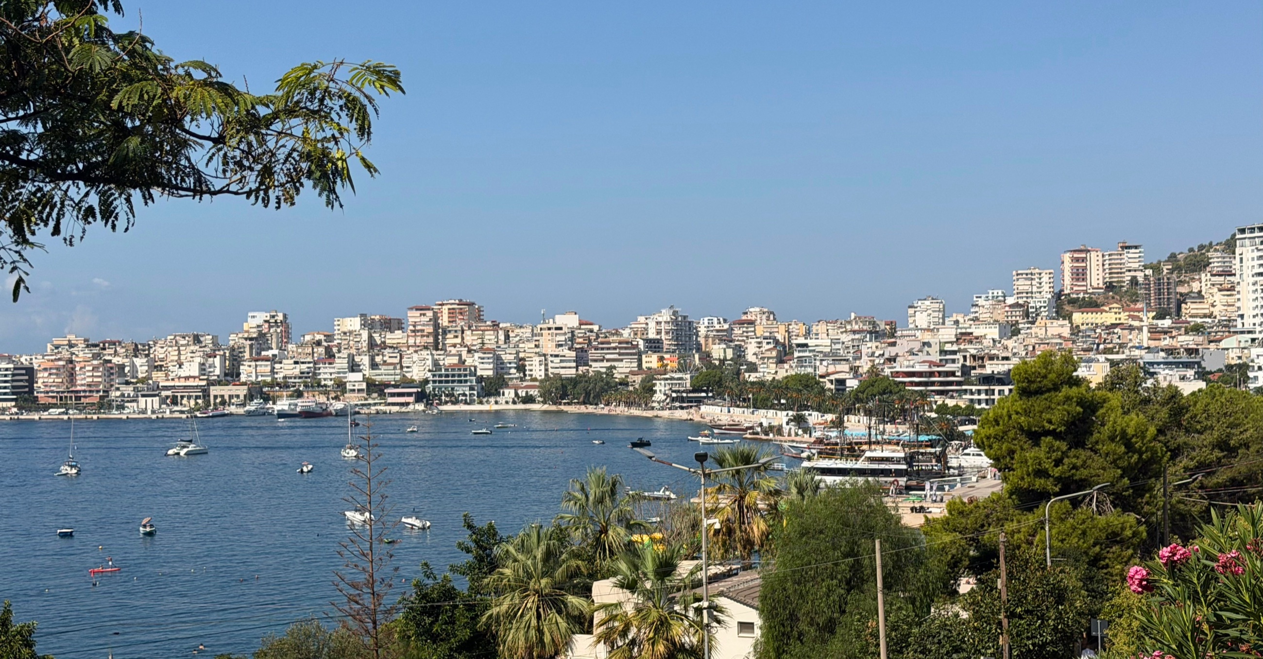 Sarandë's waterfront and hillside neighborhoods viewed across the bay in daylight.