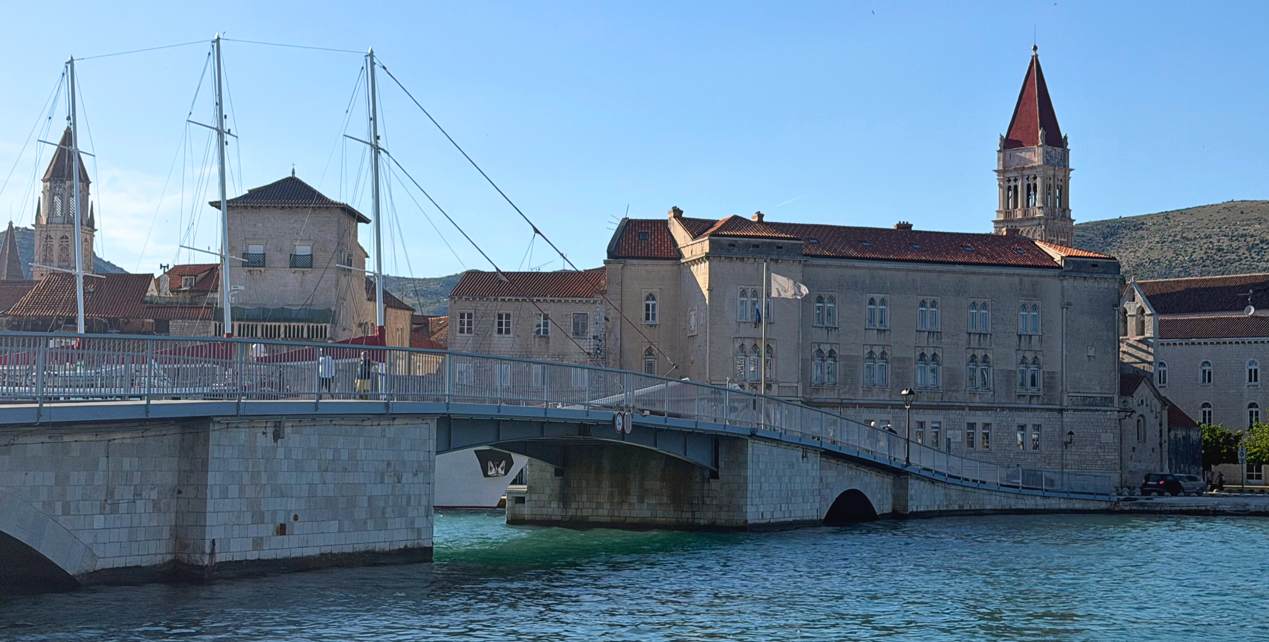 Trogir waterfront with a bridge, bell towers, stone facades, and water in the foreground.