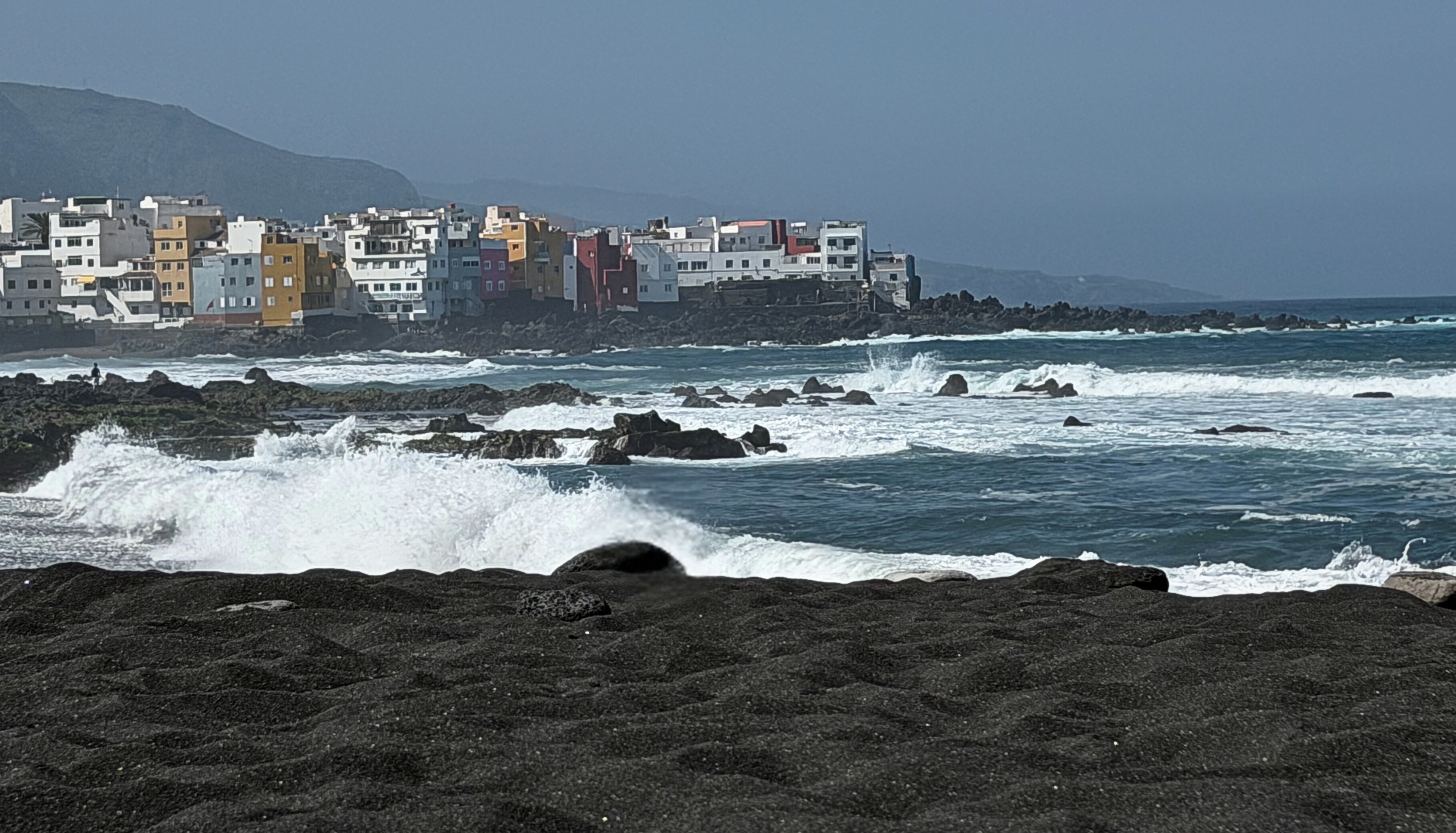 Black sand, volcanic rock, and waves at Playa Jardín in Puerto de la Cruz under hazy light.