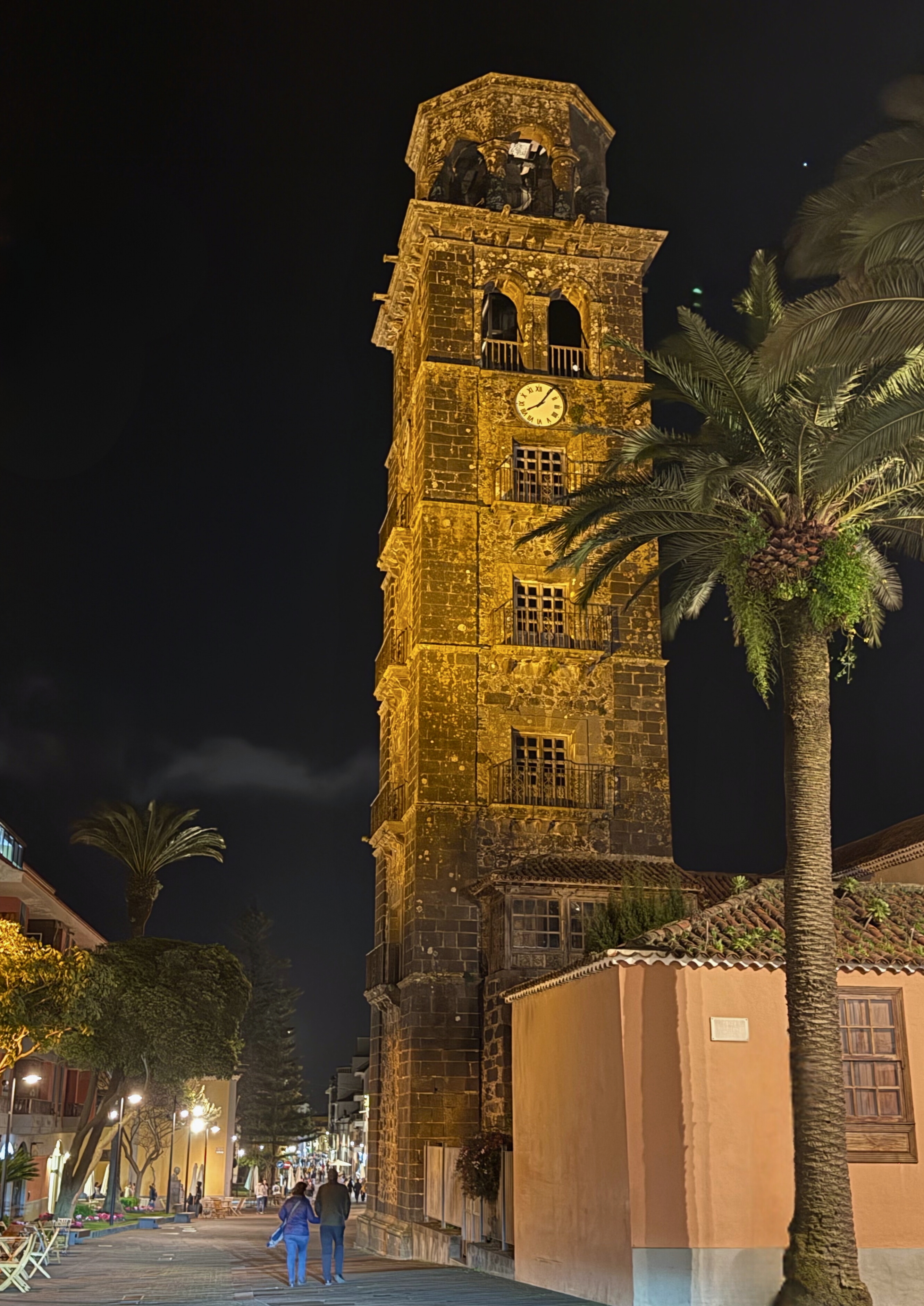 La Laguna clock tower lit at night beside a palm tree and warm stone buildings.
