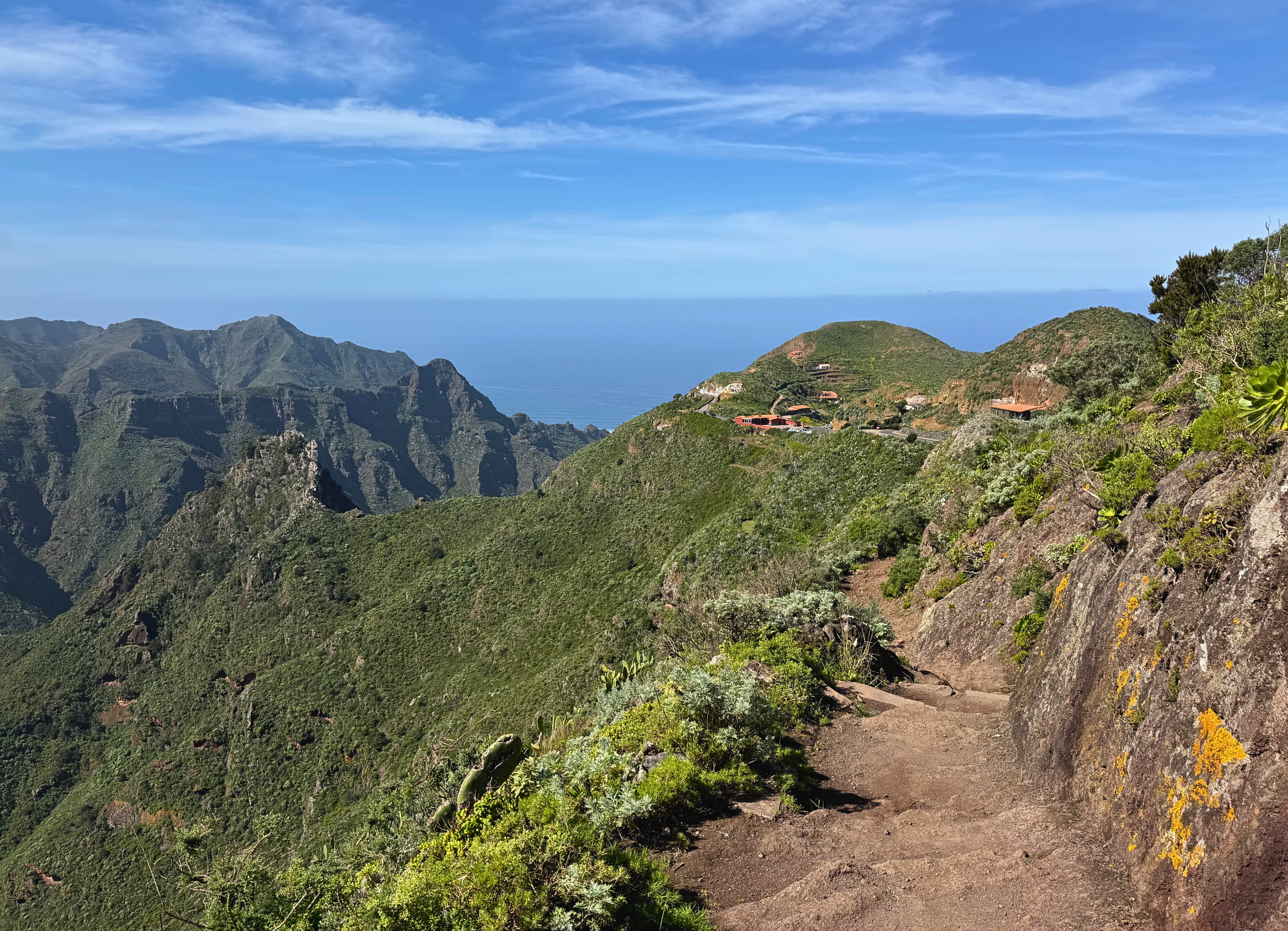 A narrow volcanic-rock trail runs through green Anaga mountains toward the sea.