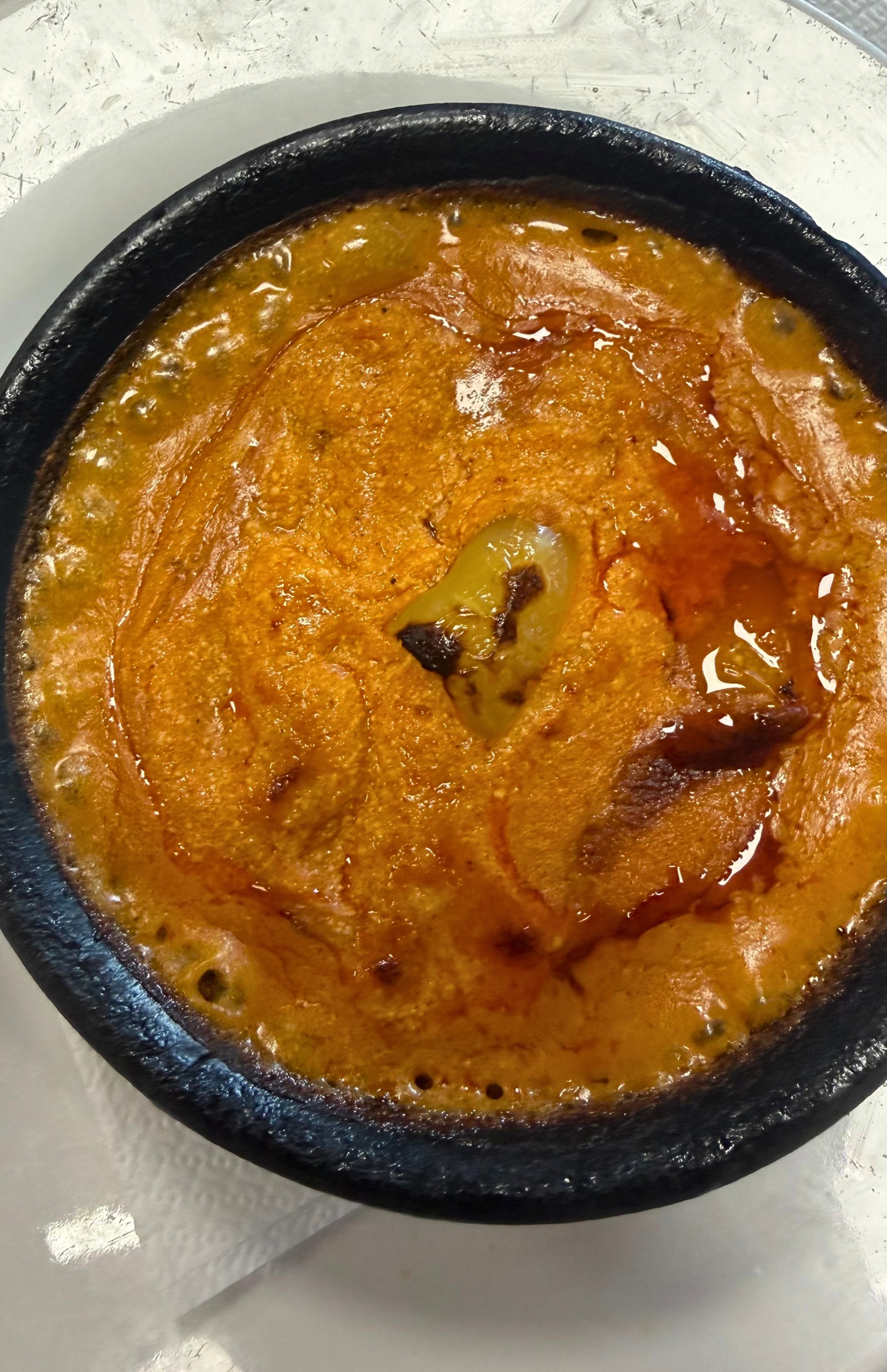 Fërgesë me speca served bubbling in a dark clay dish, with roasted peppers and cheese in a tomato-based sauce.
