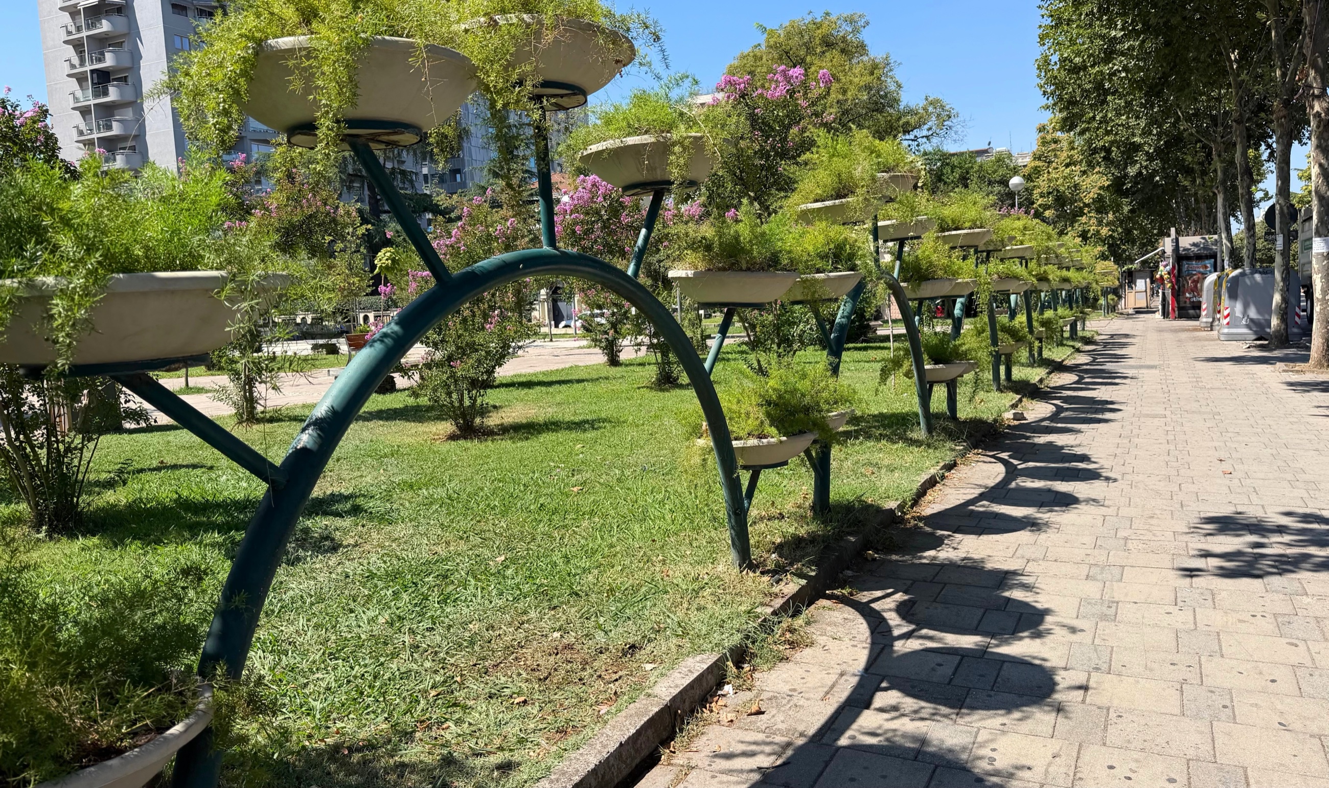 The planted walkways of Rinia Park in central Tirana, with curved metal supports holding greenery beside a paved path.