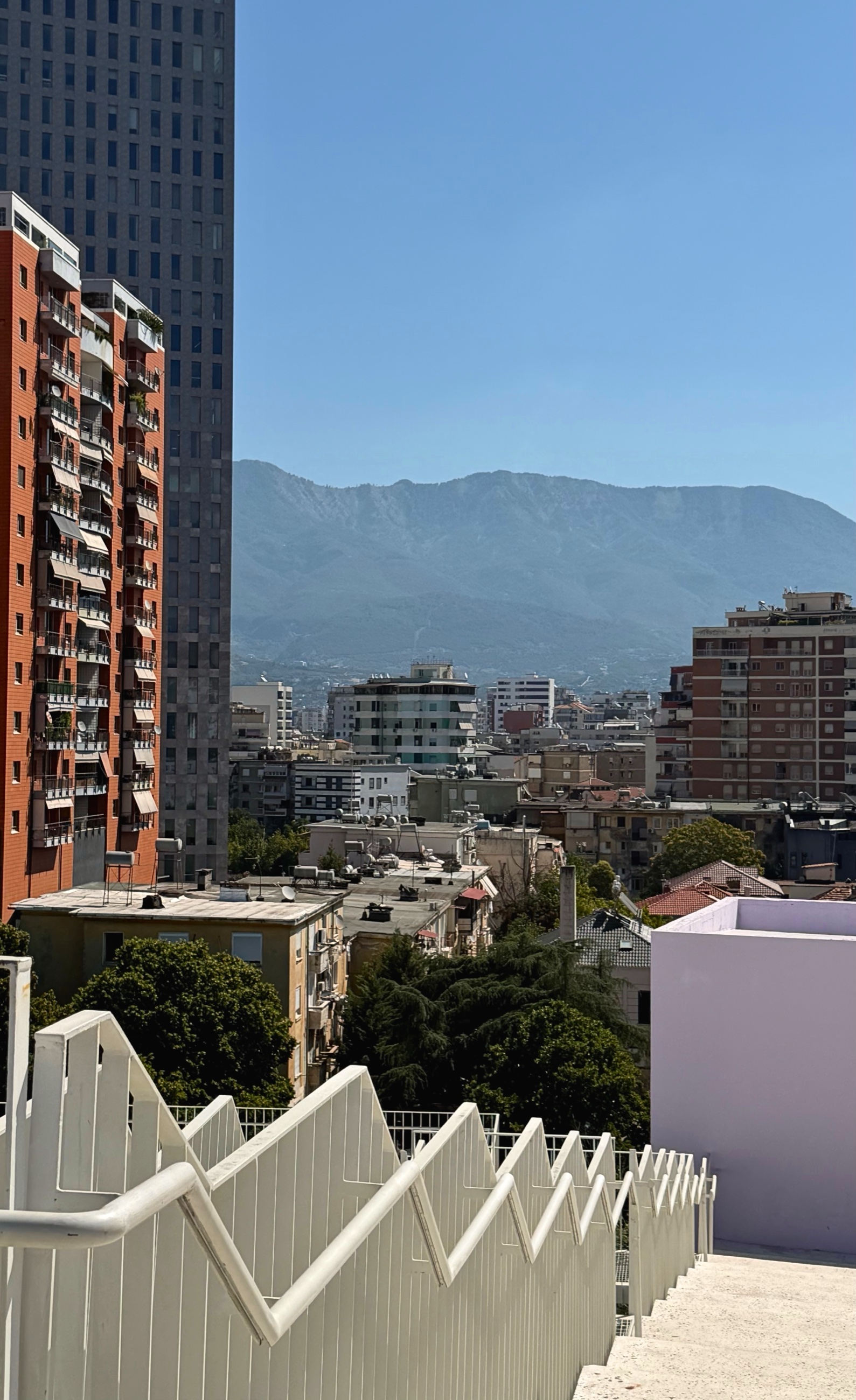 Older apartment blocks and newer towers in central Tirana rising toward Mount Dajti under a clear blue sky.