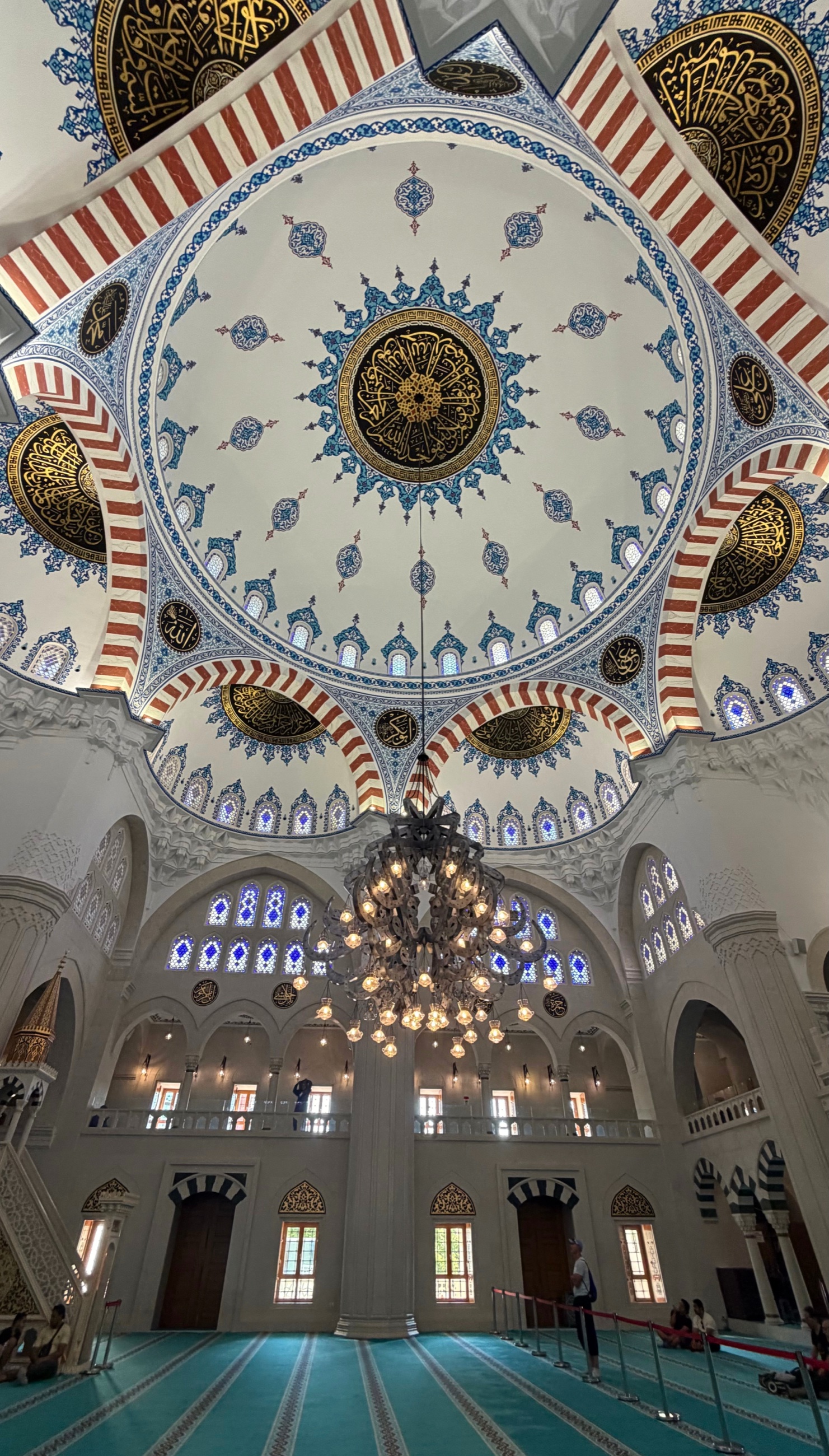 The interior of Tirana's Namazgah Mosque beneath its patterned central dome, with chandeliers, calligraphy, and blue prayer carpets.