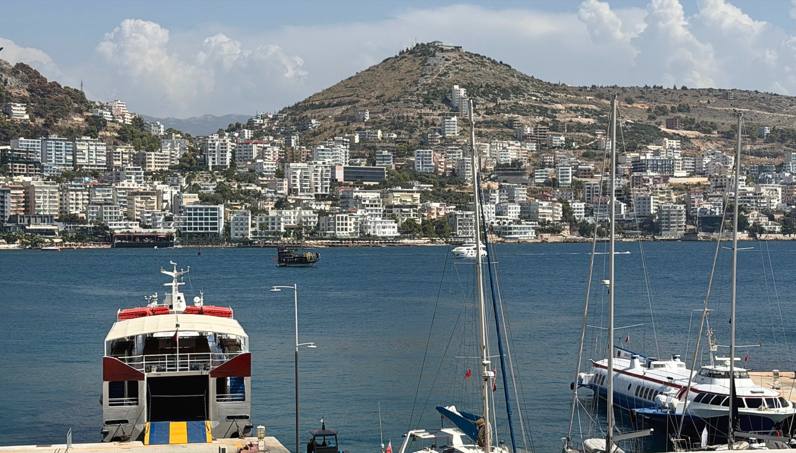Sarandë harbor with ferries and sailboats below the hillside city.