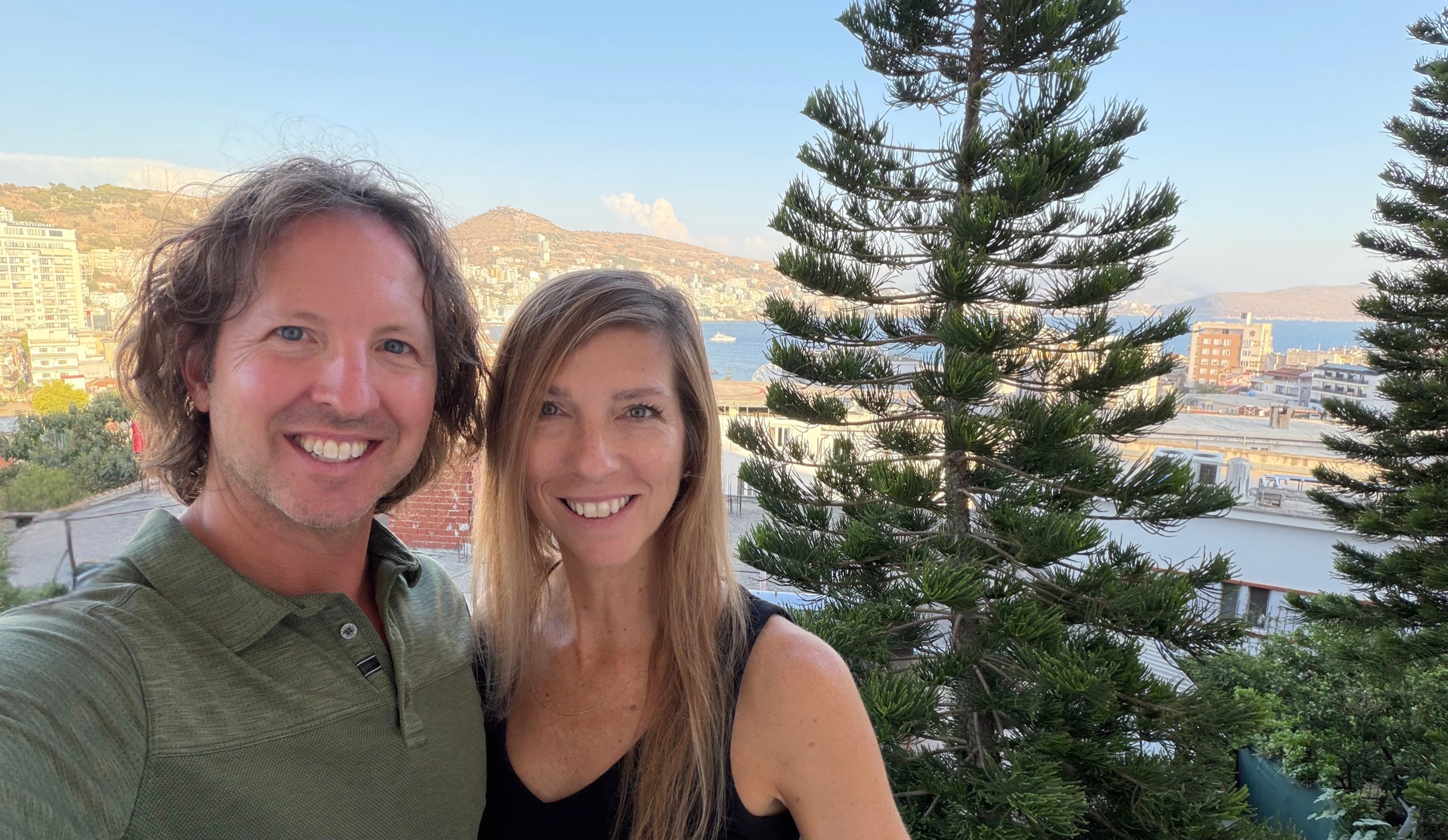 Sam and Stephanie on the balcony of their Sarandë apartment with the hillside city and Ionian Sea behind them.