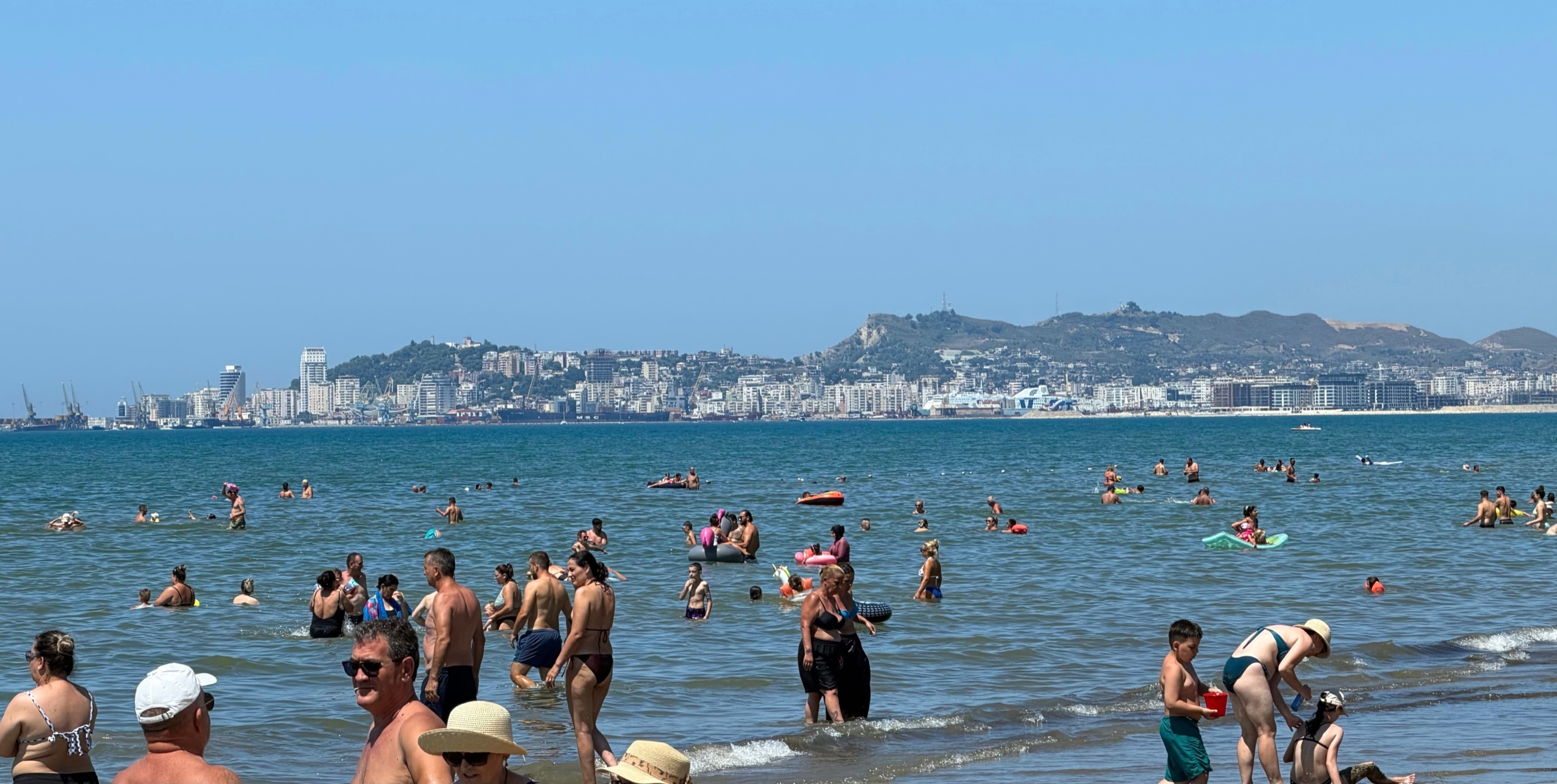Crowds swimming in the warm, shallow water at Durrës Beach, with the city stretching around the bay.