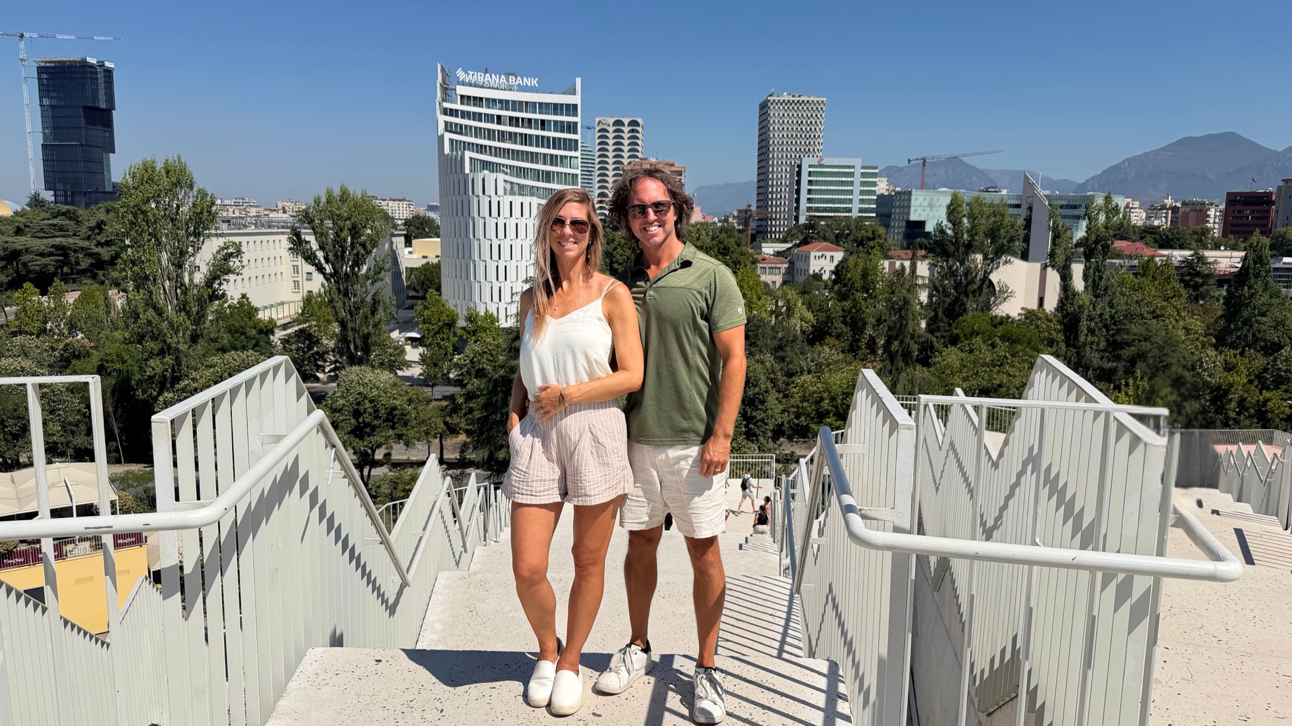 Sam and Stephanie standing together on the rebuilt terraces of the Pyramid of Tirana, with the city skyline behind them.