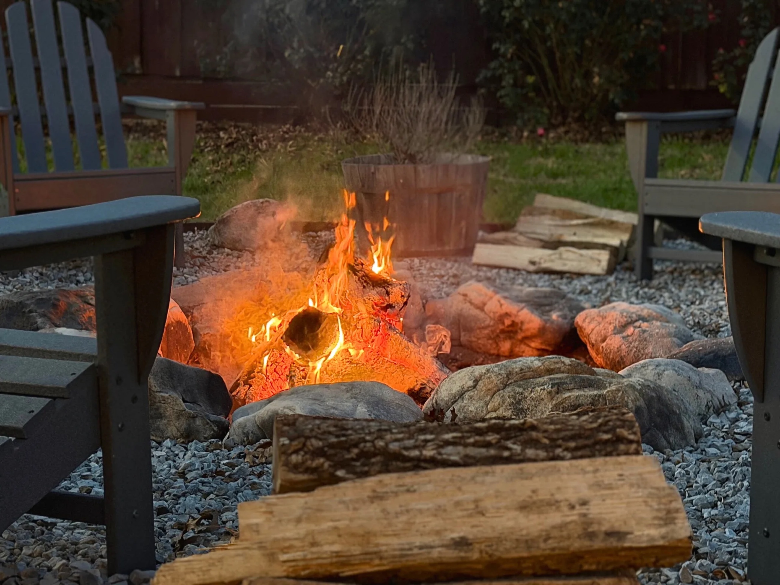 A fire pit burning in a backyard at dusk, surrounded by Adirondack chairs.