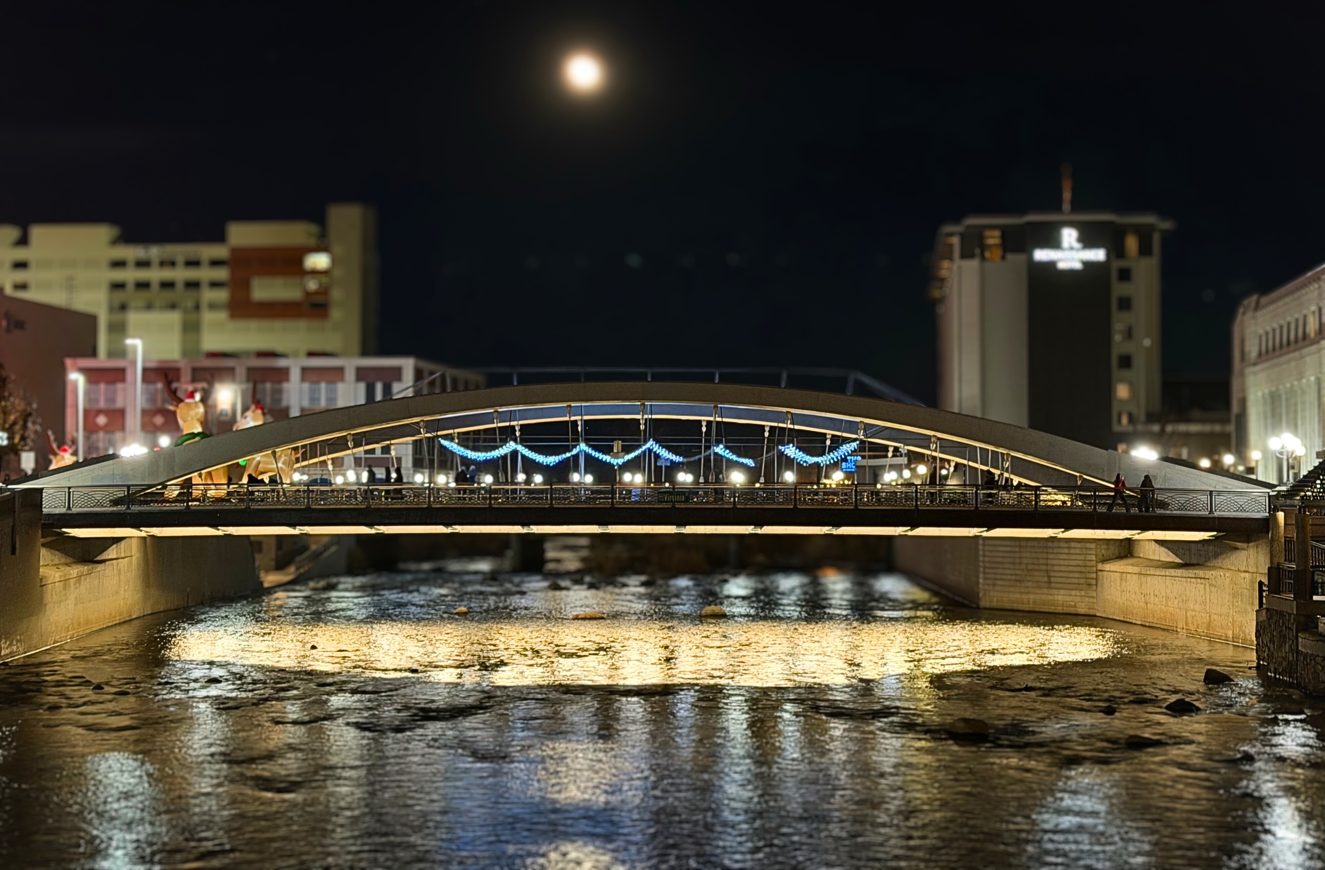 A bridge decorated with holiday lights reflected in the Truckee River at night.
